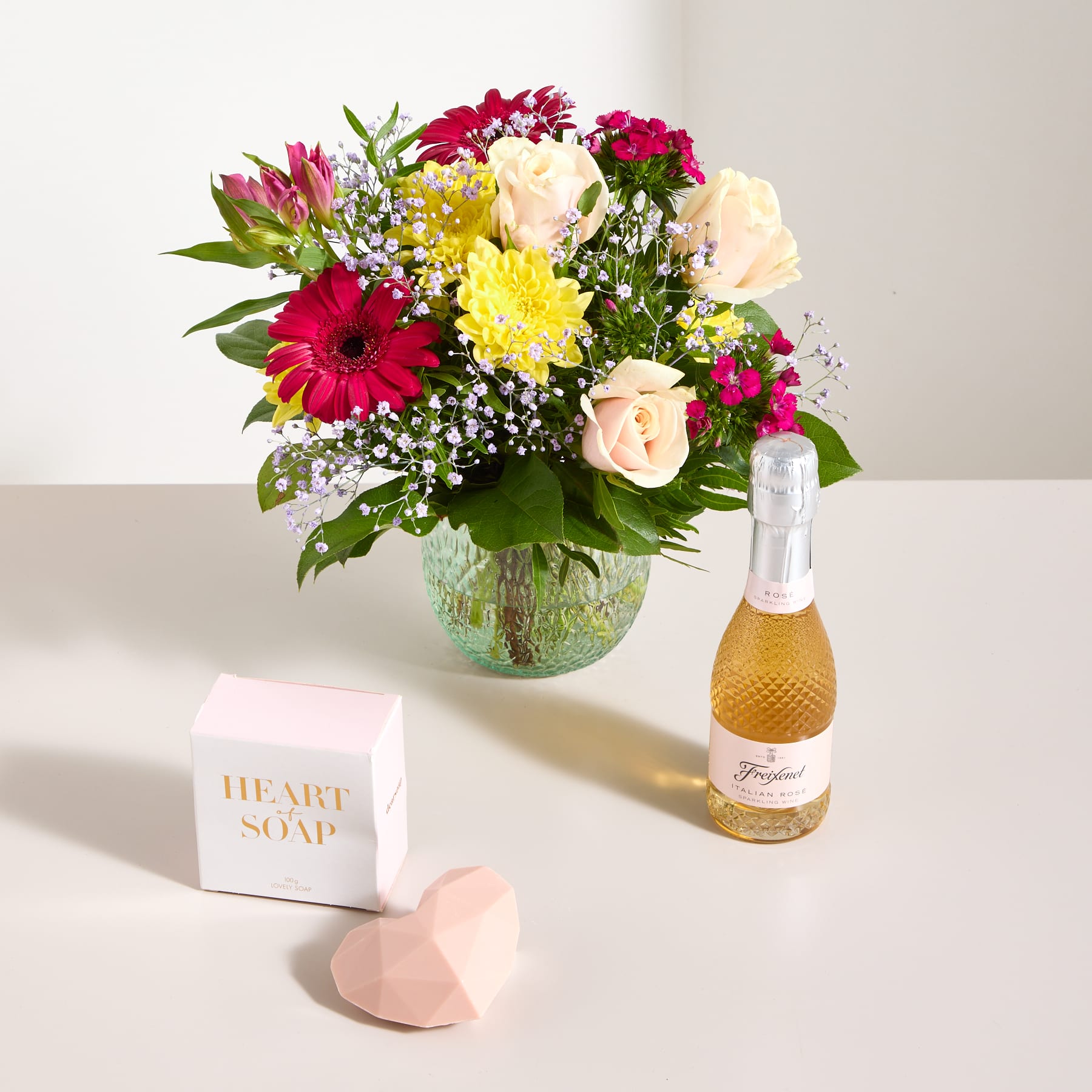 Ein farbenfroher Strauß bunter Blumen, darunter Rosen, Gerbera und andere Blüten, steht neben einer Flasche Champagner und einer herzförmigen Seife auf einer hellen Oberfläche.