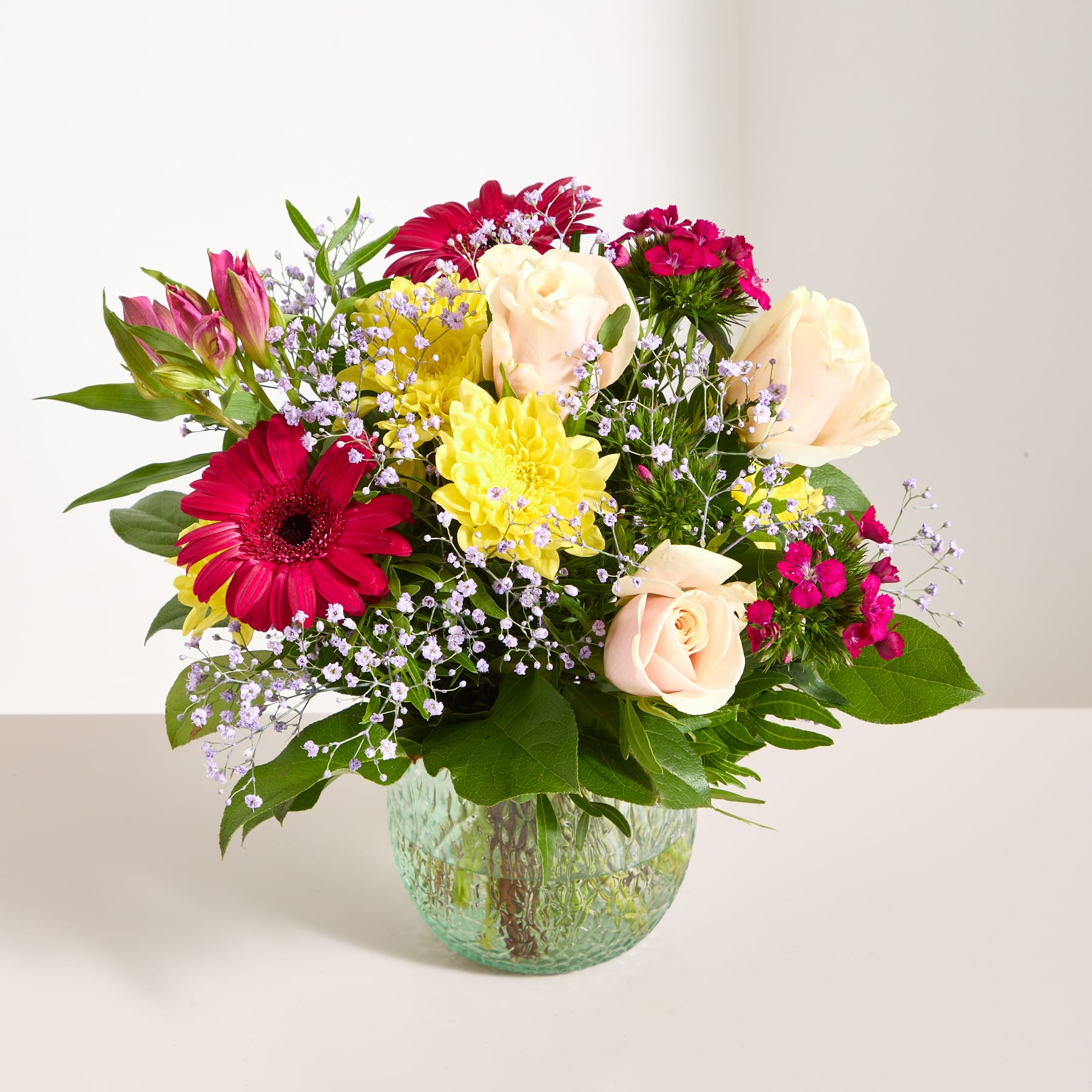Ein lebendiger Strauß verschiedener bunter Blumen, darunter Rosen, Gerbera und andere Blüten, arrangiert in einer Glasvase vor einem schlichten Hintergrund.
