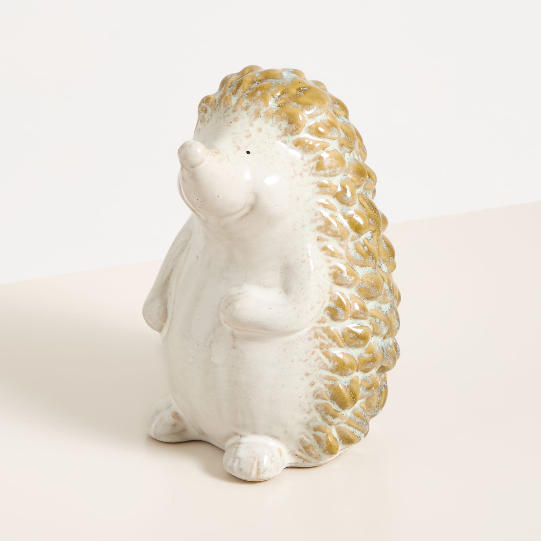 Eine kleine, weiße Keramikfigur eines igel- oder stachelschweinähnlichen Wesens mit strukturiertem, goldfarbenem Panzer steht auf einer schlichten, hellen Oberfläche.