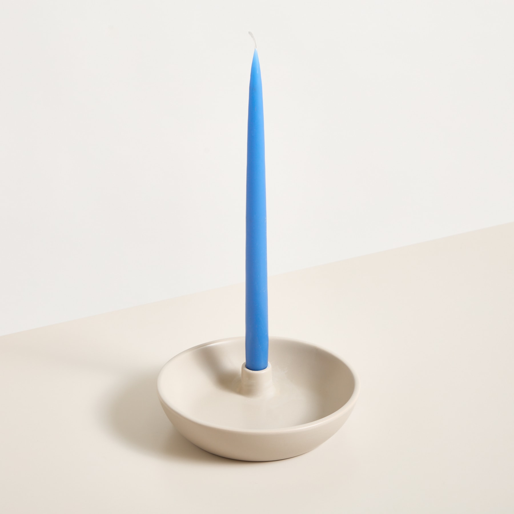Eine blaue Kerze steht aufrecht in einer weißen Keramikschale vor einem schlichten, hellen Hintergrund.