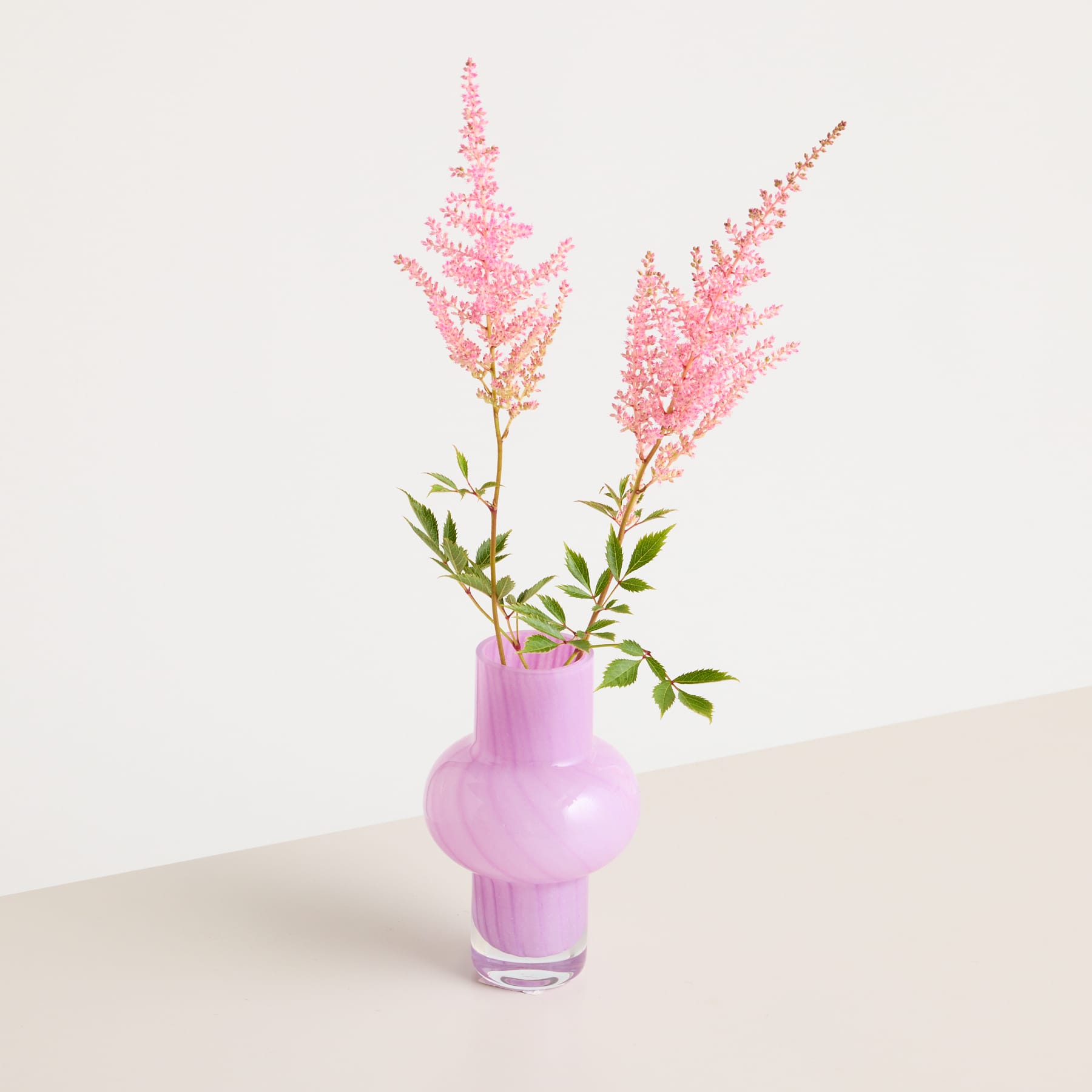 Ein zarter Strauß rosa Astilbenblüten steht in einer hellvioletten Vase vor einem schlichten, hellen Hintergrund.