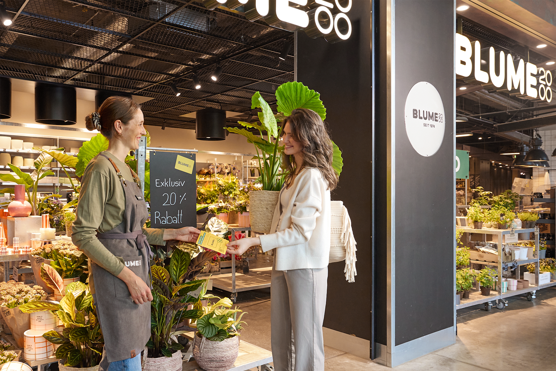 Zwei Frauen unterhalten sich in einem lebhaften, mit Pflanzen gefüllten Ladenlokal. Im Hintergrund sind verschiedene Produkte und Schilder zu sehen.
