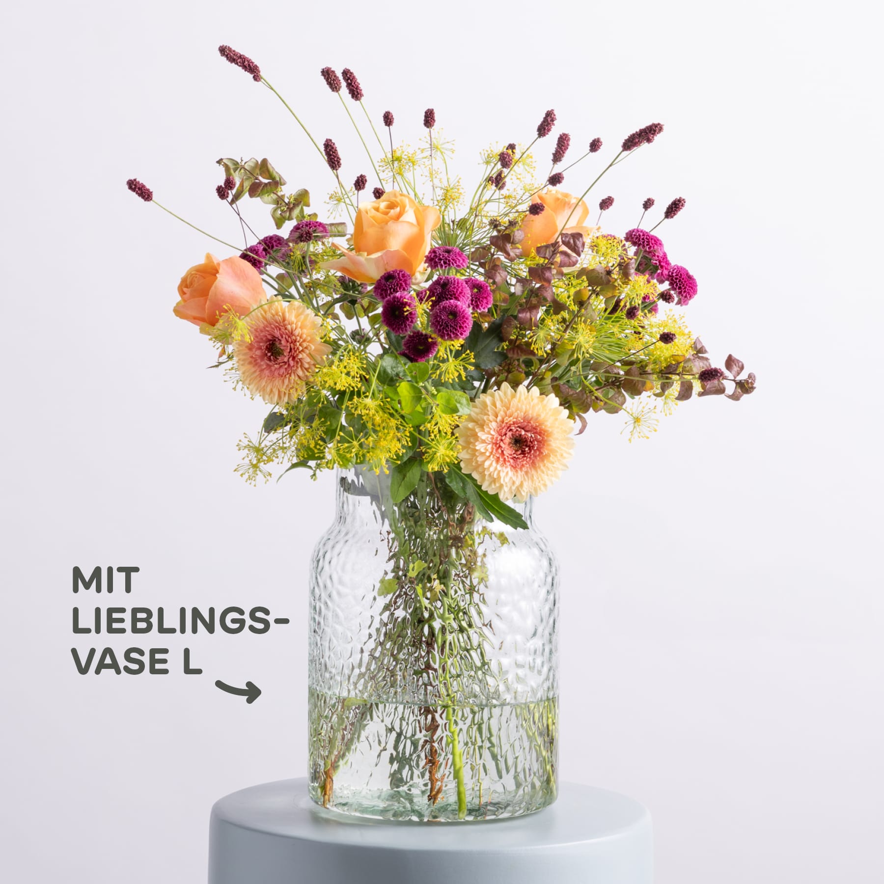 Ein lebendiger Strauß bunter Blumen, darunter Gänseblümchen, Rosen und andere Blüten, wird in einer Glasvase auf einem schlichten weißen Hintergrund präsentiert.