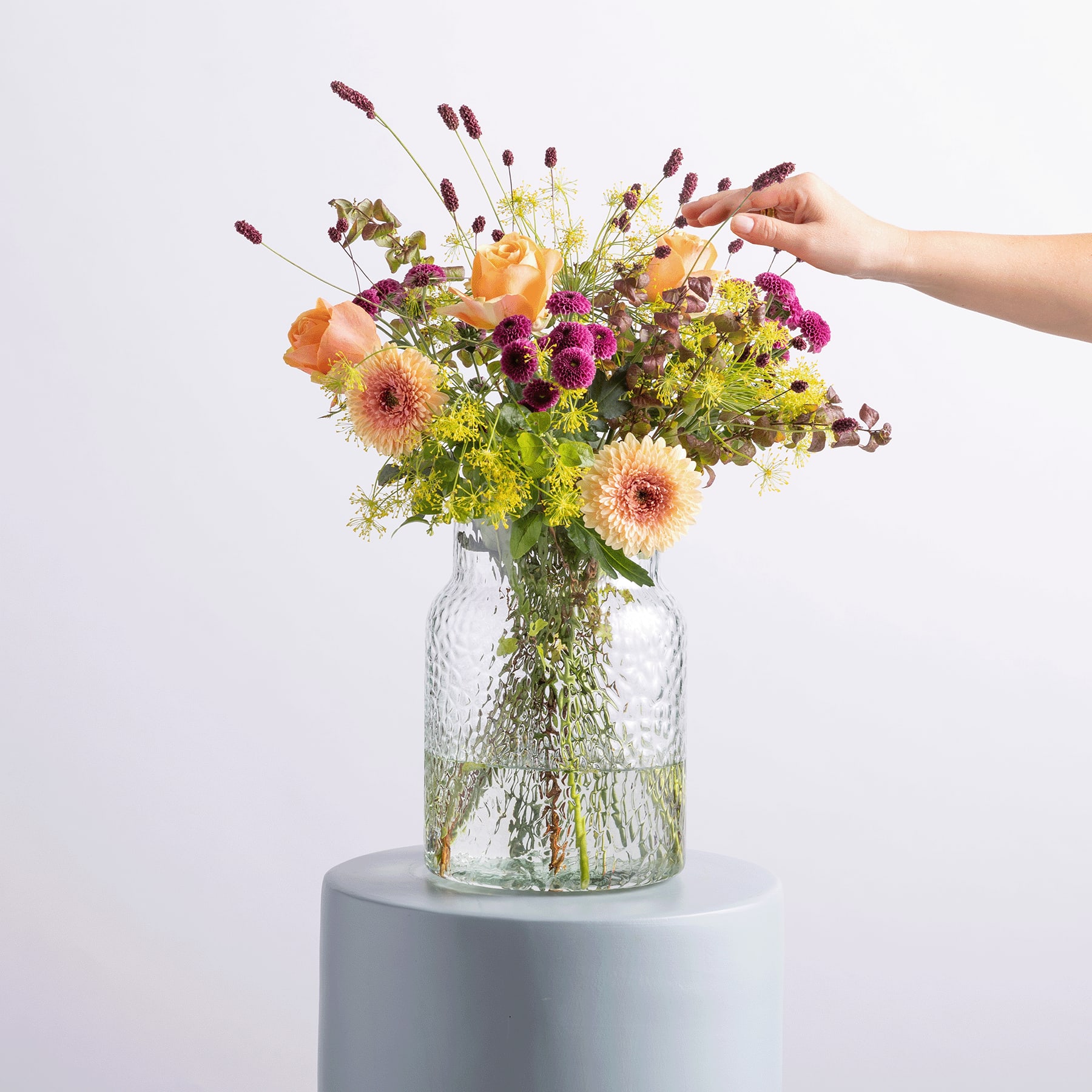 Ein lebendiger Strauß bunter Blumen, darunter Gänseblümchen, Chrysanthemen und andere Blüten, wird in einer Glasvase vor einem schlichten weißen Hintergrund präsentiert.