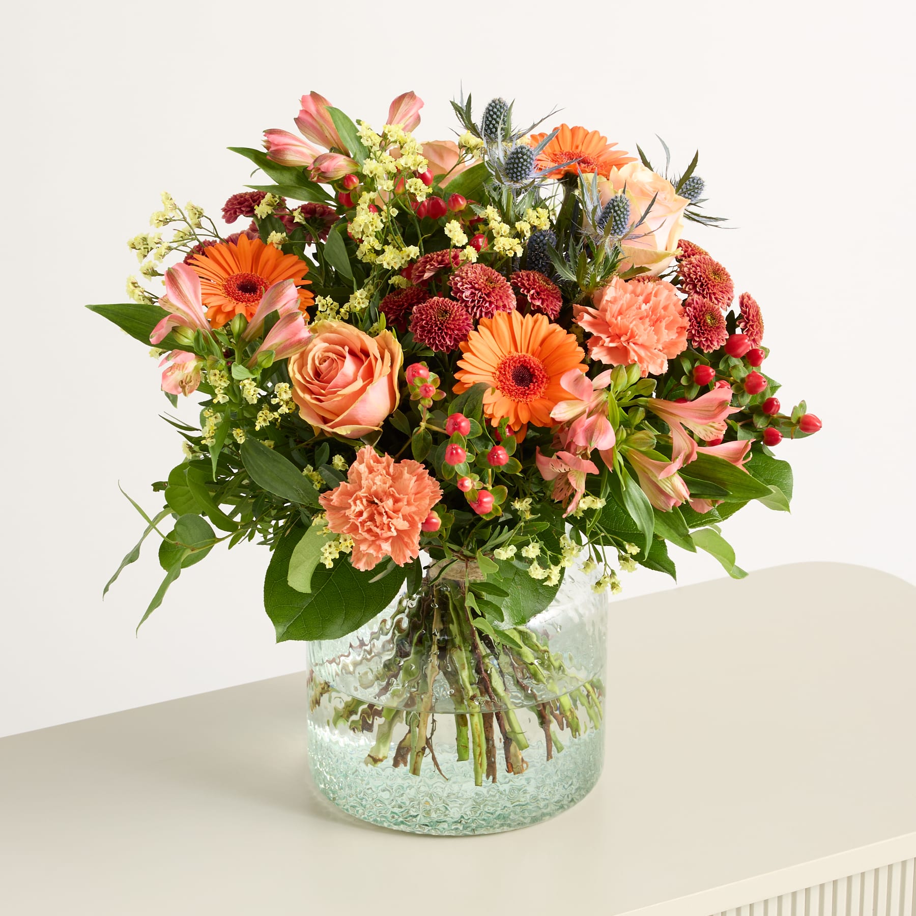 Ein farbenfroher Strauß verschiedener Blumen, darunter Rosen, Gerbera und Nelken, arrangiert in einer Glasvase auf einem schlichten Hintergrund.