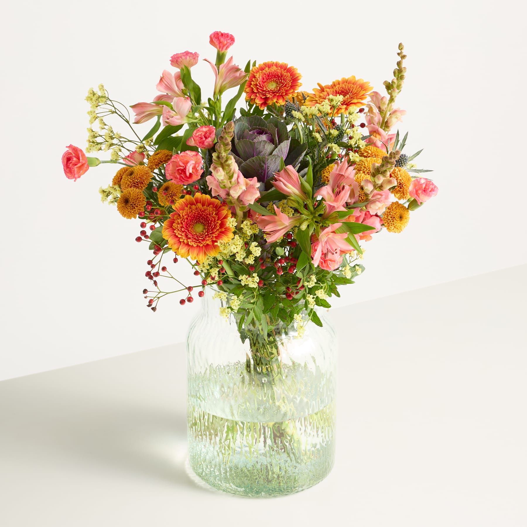 Ein lebendiger Strauß bunter Blumen, darunter Sonnenblumen, Nelken und andere Blüten, wird in einer klaren Glasvase vor einem schlichten weißen Hintergrund präsentiert.