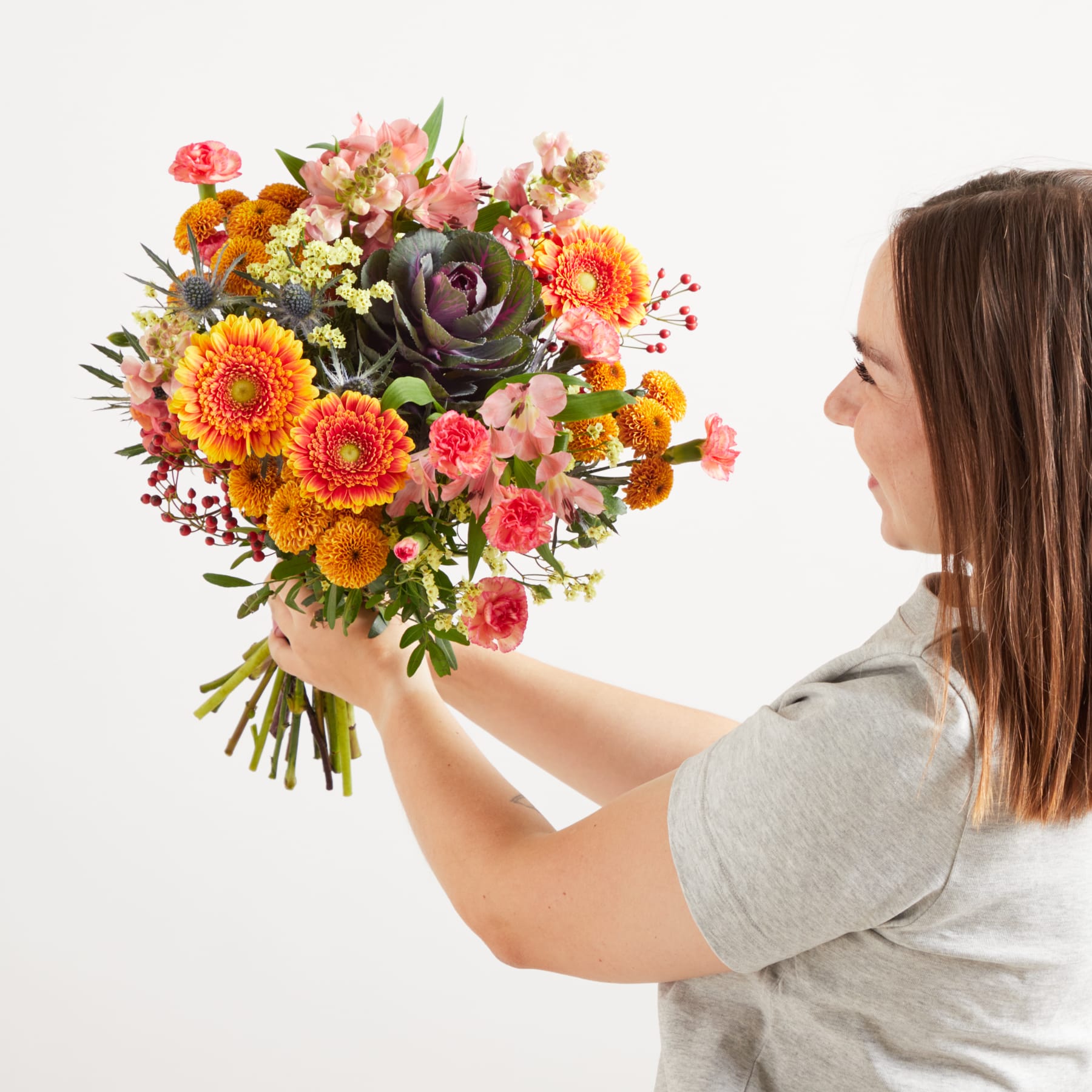 Eine Frau hält vor einem schlichten weißen Hintergrund einen leuchtenden Strauß bunter Blumen, darunter Sonnenblumen, Rosen und andere herbstlich inspirierte Blüten.