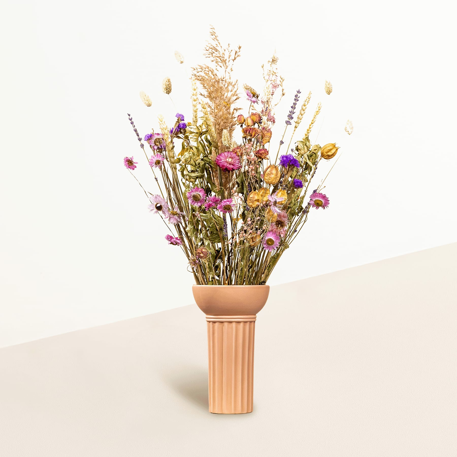 Ein Strauß leuchtender Trockenblumen und Gräser in einer Terrakotta-Vase steht vor einem schlichten, hellen Hintergrund.
