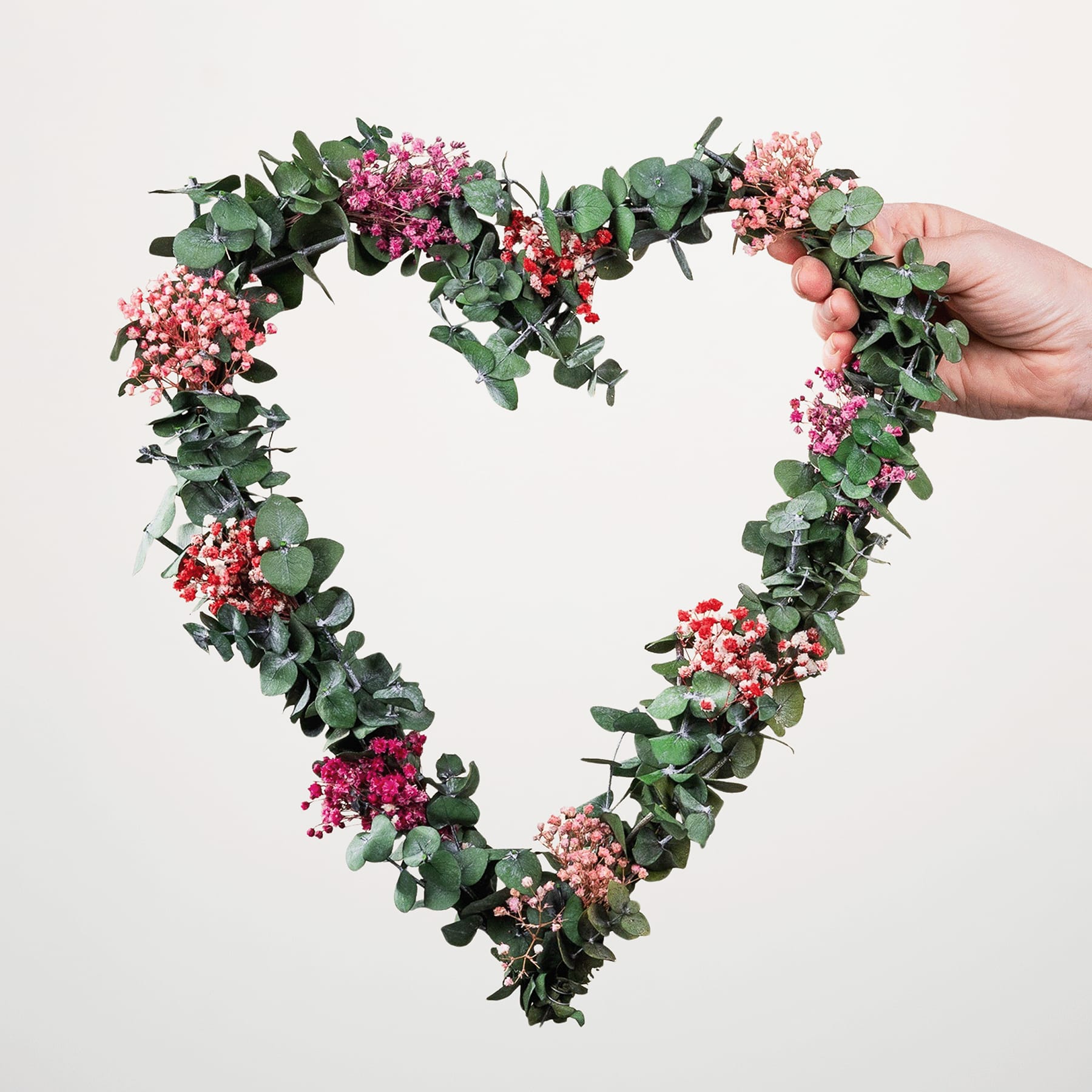 Ein herzförmiger Kranz aus üppigen grünen Blättern und leuchtend rosa Blüten wird vor einem schlichten weißen Hintergrund hochgehalten.