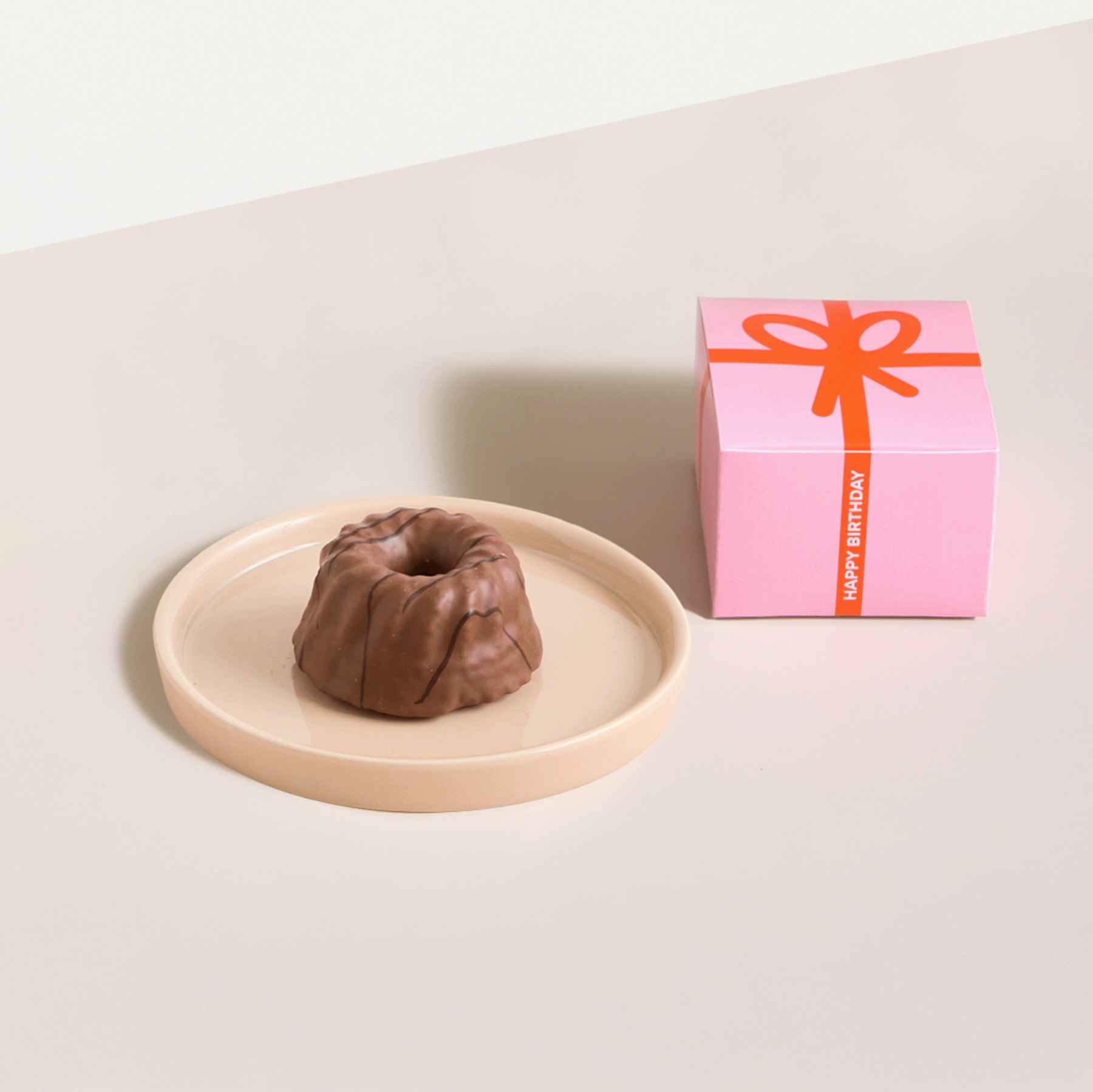 Eine rosa Geschenkbox mit orangefarbener Schleife steht neben einem runden Schokoladendessert auf einem hellen Teller.