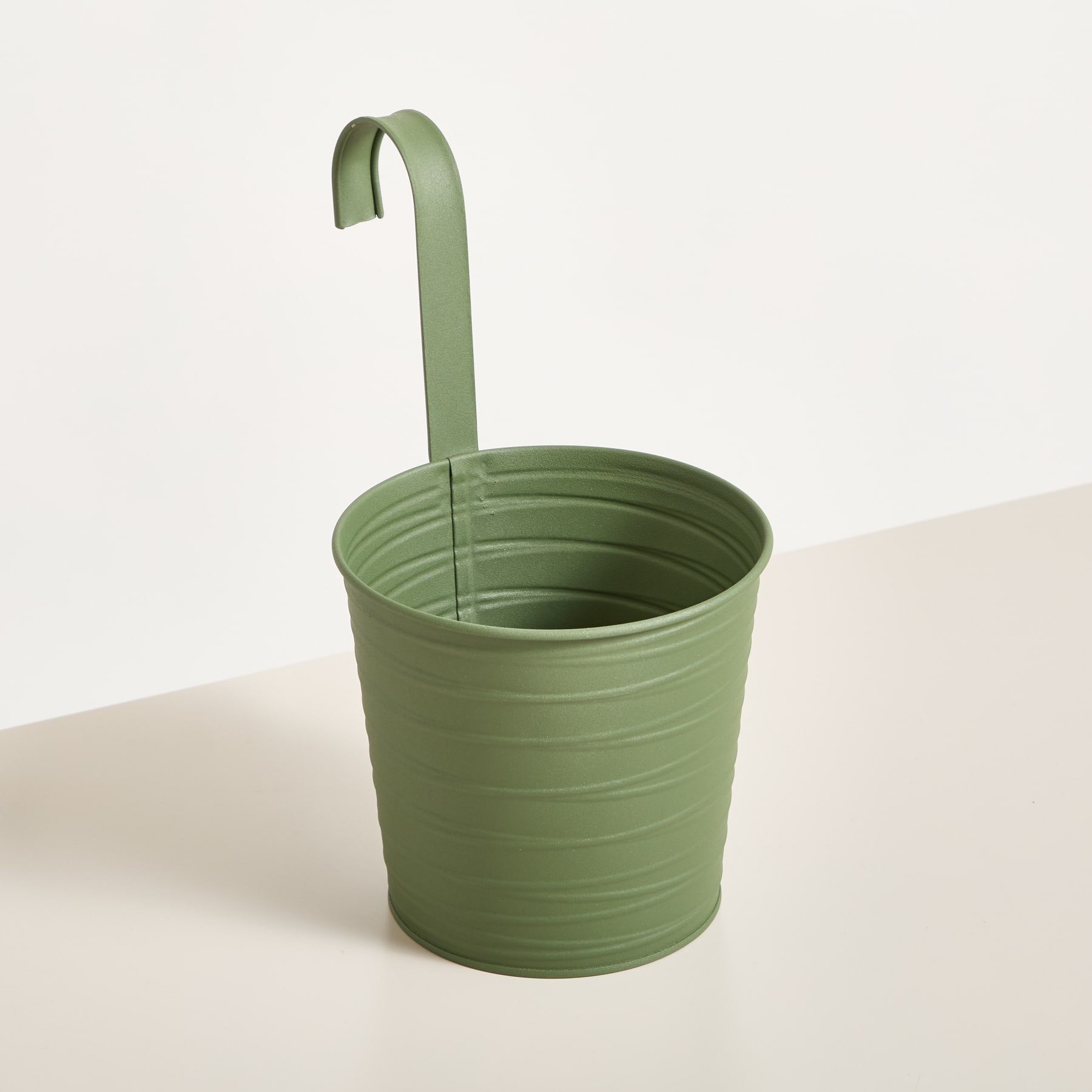 Ein grüner Blumentopf oder Behälter aus Metall mit einem gebogenen Griff, der auf einer hellen Oberfläche vor einem schlichten Hintergrund steht.