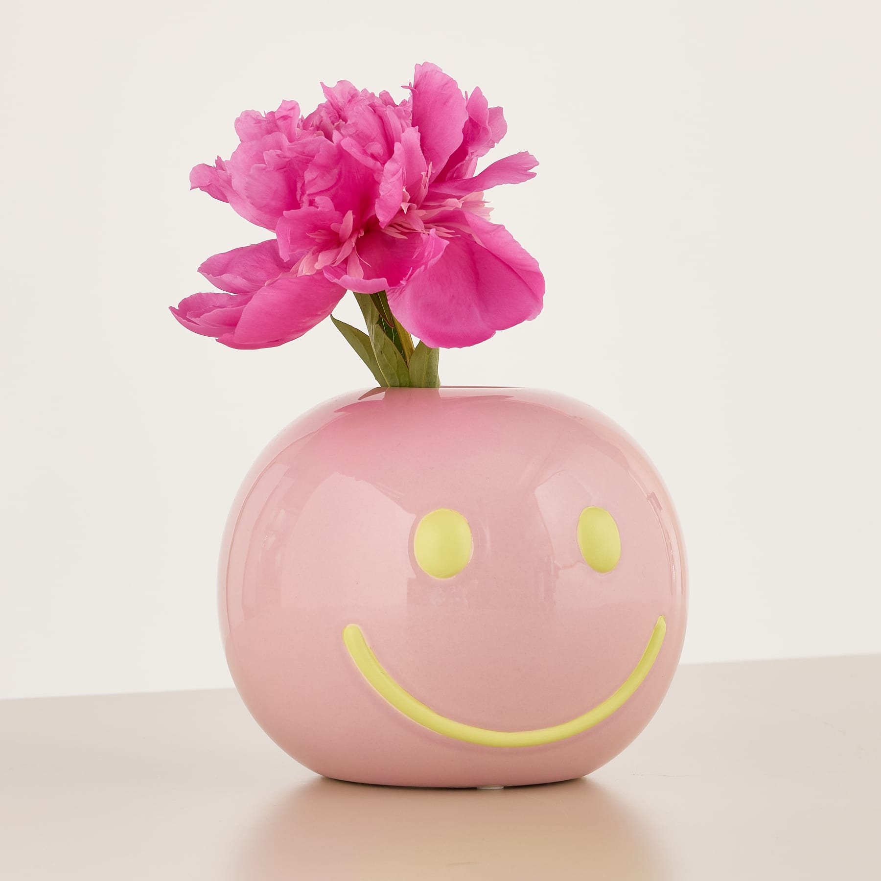 Eine leuchtend rosa Pfingstrosenblüte wird in einer rosa Keramikvase mit Smiley-Design vor einem neutralen Hintergrund präsentiert.