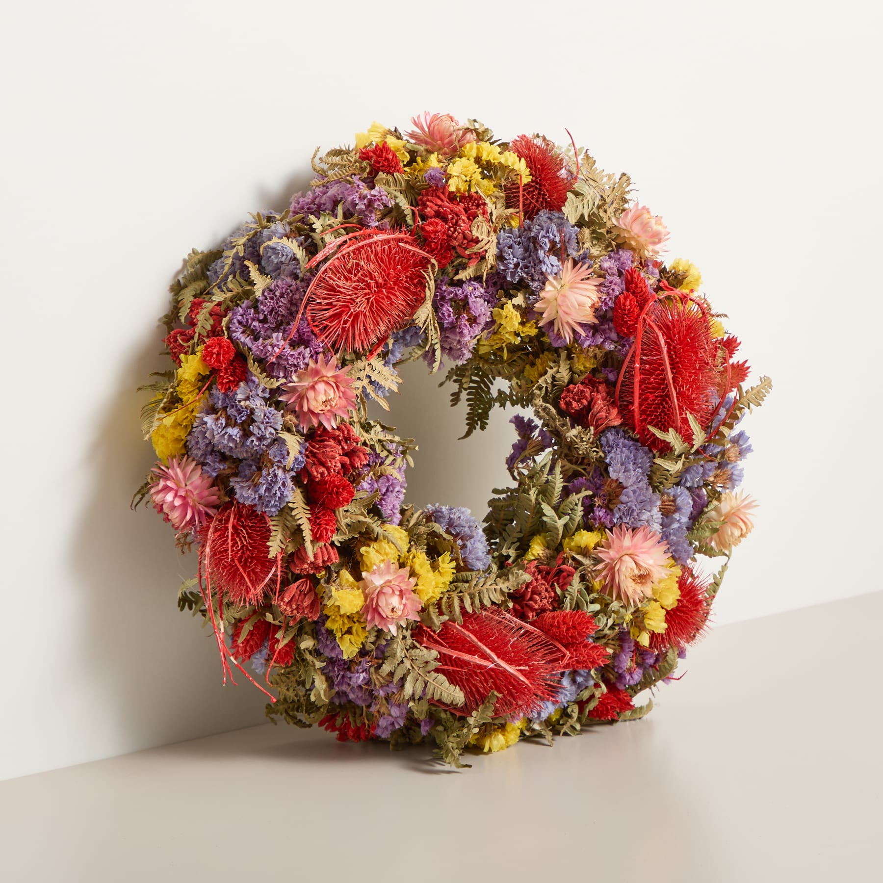 Ein leuchtender, runder Kranz aus verschiedenen bunten Blumen, darunter rote, gelbe und violette Blüten, liegt auf einer Holzoberfläche vor einem schlichten Hintergrund.