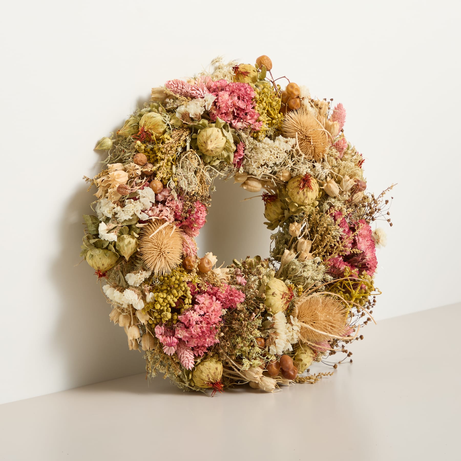 Ein farbenfroher Blumenkranz aus verschiedenen getrockneten Blumen und Blättern, gehalten in einer behandschuhten Hand vor einem schlichten weißen Hintergrund.