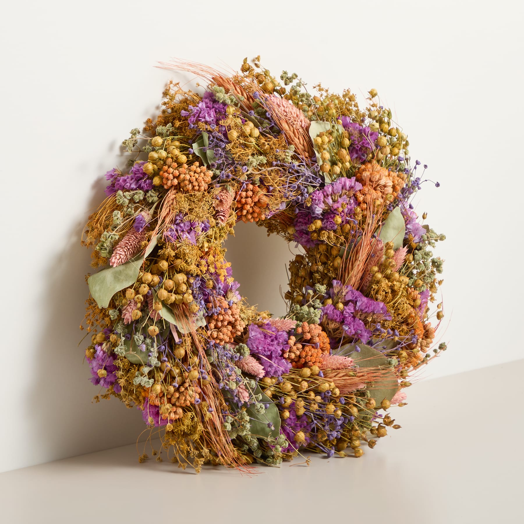 Vor einem schlichten weißen Hintergrund ist ein leuchtender, runder Kranz aus verschiedenen getrockneten Blumen und Blättern in Gelb-, Orange- und Lilatönen zu sehen.