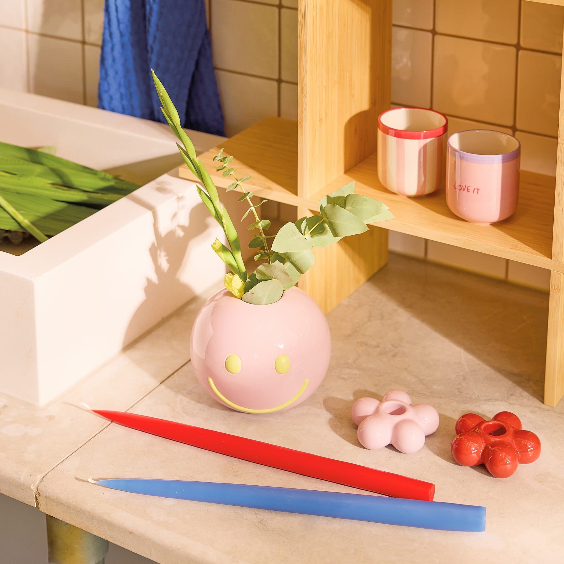 Eine leuchtend rosa Pfingstrosenblüte wird in einer skurrilen rosa Keramikvase mit Smiley-Design vor einem schlichten, hellen Hintergrund präsentiert.