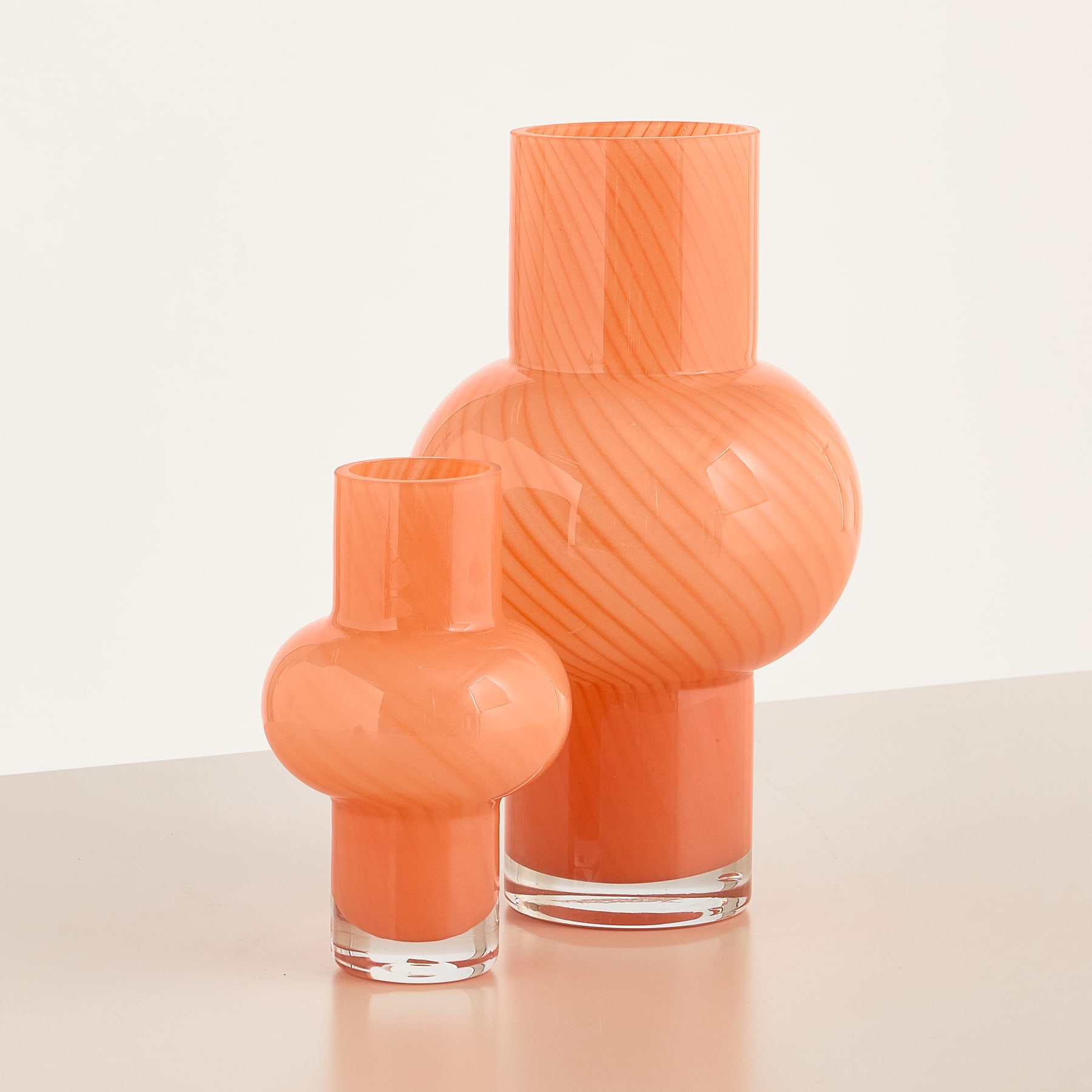 Das Bild zeigt zwei leuchtend orangefarbene Glasvasen oder -gefäße unterschiedlicher Größe, die auf einer hellen Oberfläche vor einem schlichten Hintergrund platziert sind.