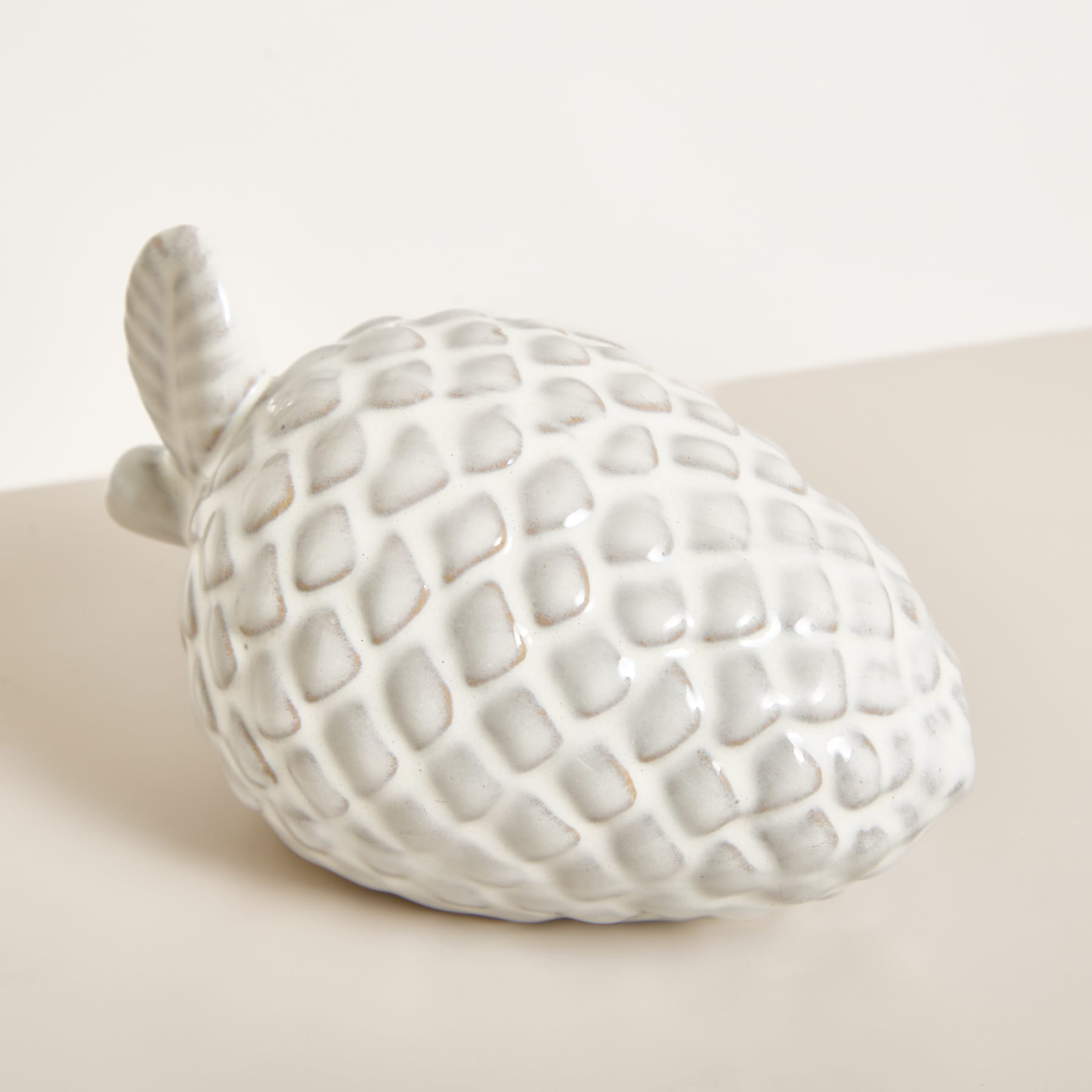 Ein weißes, strukturiertes Keramikobjekt, das einer Ananas oder Artischocke ähnelt, liegt auf einer hellen Oberfläche vor einem schlichten Hintergrund.