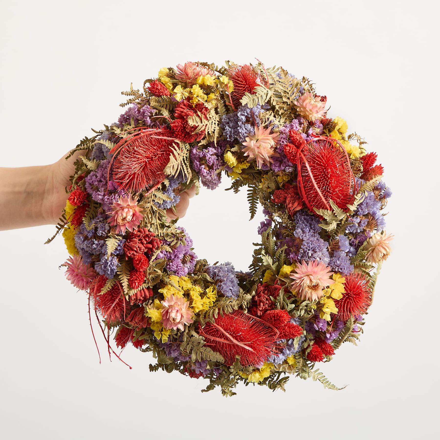 Ein lebendiger und farbenfroher Kranz aus verschiedenen Blumen, darunter rote, gelbe und violette Blüten, steht auf einem Holzregal vor einem schlichten Hintergrund.
