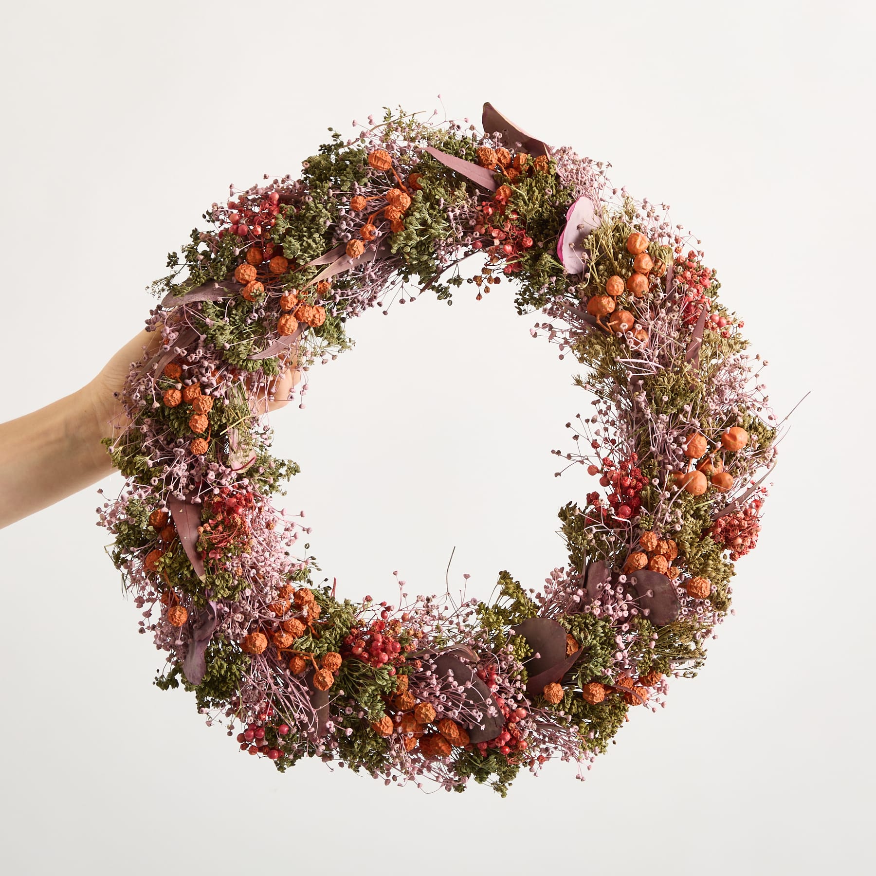 Ein farbenfroher, runder Kranz aus verschiedenen getrockneten Blumen, Beeren und anderen natürlichen Elementen wird vor einem schlichten weißen Hintergrund in den Händen einer Person gehalten.