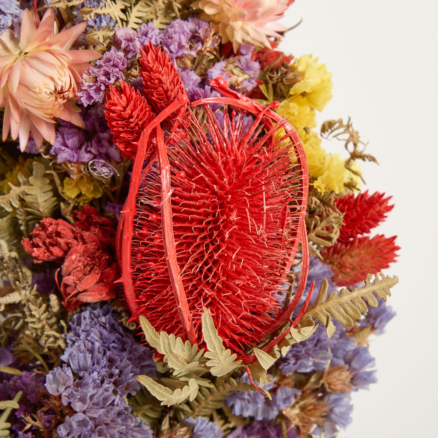 Ein lebendiger Strauß aus verschiedenen bunten Blumen, darunter auffällige rote Protea-Blüten, umgeben von einer Mischung aus violetten, gelben und orangefarbenen Farbtönen, die ein optisch ansprechendes und harmonisches Arrangement ergeben.