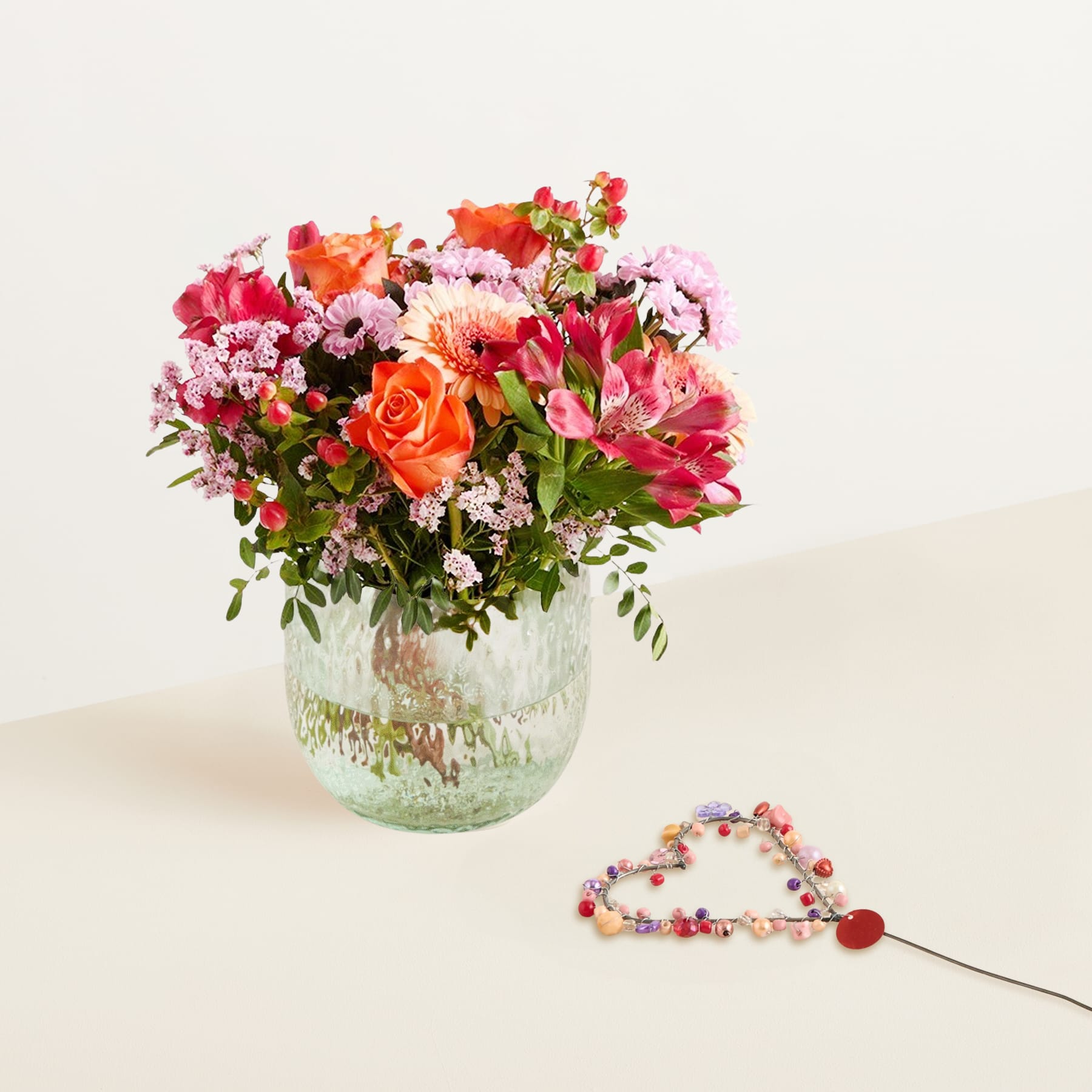 Ein farbenfroher Strauß bunter Blumen, darunter Rosen und Nelken, steht in einer Glasvase vor einem schlichten, hellen Hintergrund. Im Vordergrund ist eine Perlenkette oder ein Perlenarmband zu sehen.