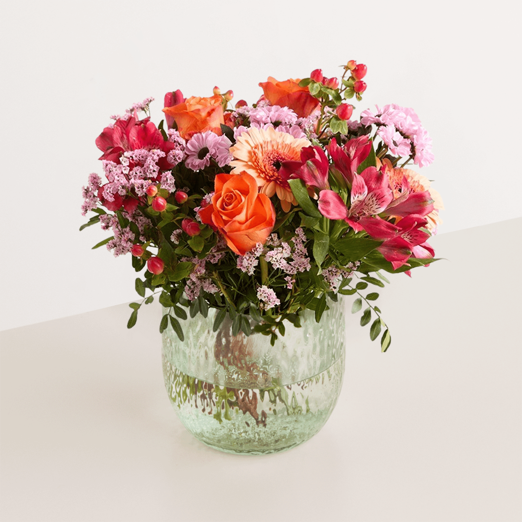 Ein farbenfroher Strauß verschiedener bunter Blumen, darunter Rosen, Gerbera und andere Blüten, wird in einer Glasvase vor einem schlichten weißen Hintergrund präsentiert.