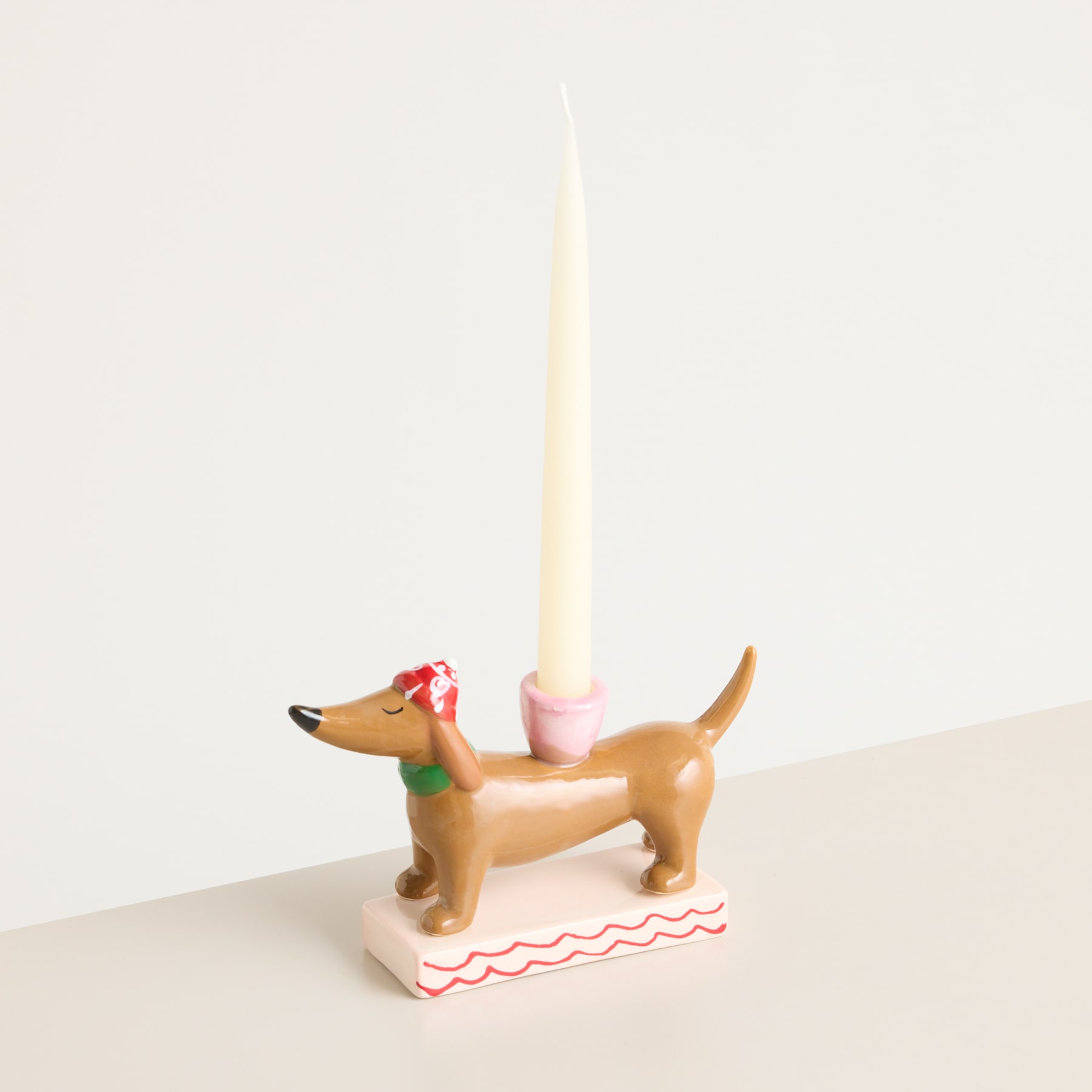 Ein kleiner, brauner Kerzenhalter in Hundeform mit einer roten Schleife auf dem Kopf, der auf einer weißen Fläche vor einem schlichten Hintergrund steht.