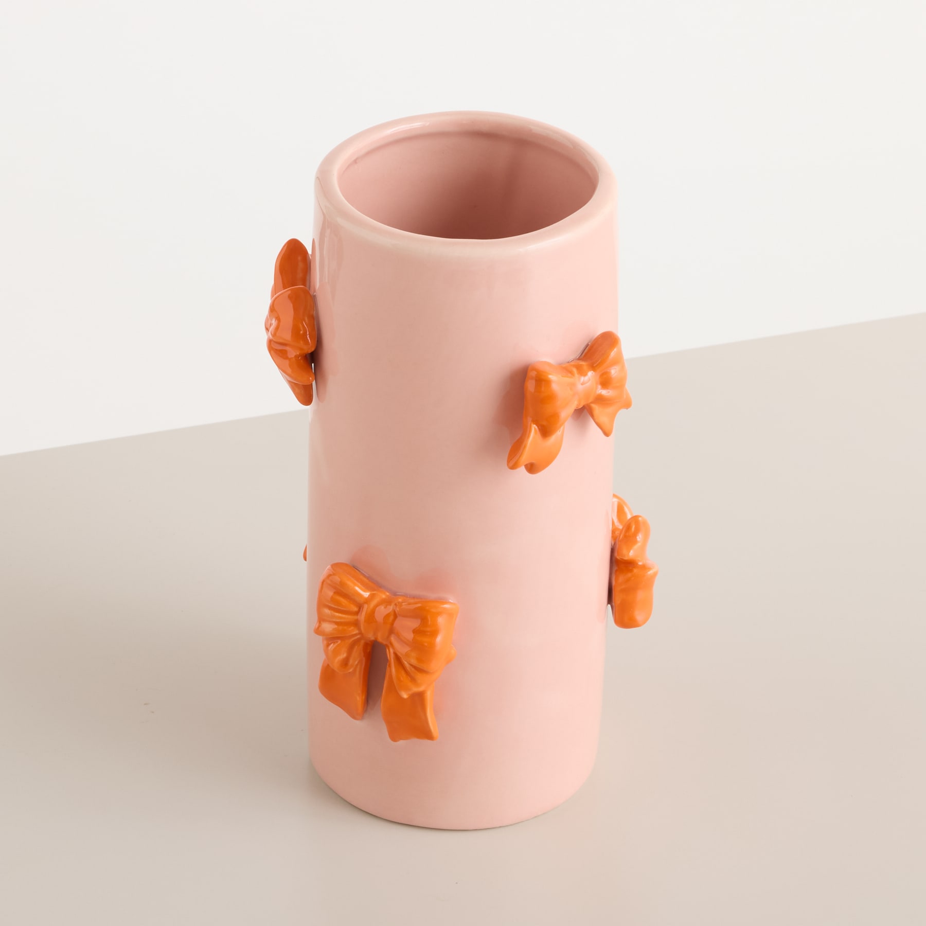 Eine rosafarbene Keramikvase mit orangefarbenen Schleifenverzierungen steht auf einer hellen Oberfläche vor einem schlichten weißen Hintergrund.