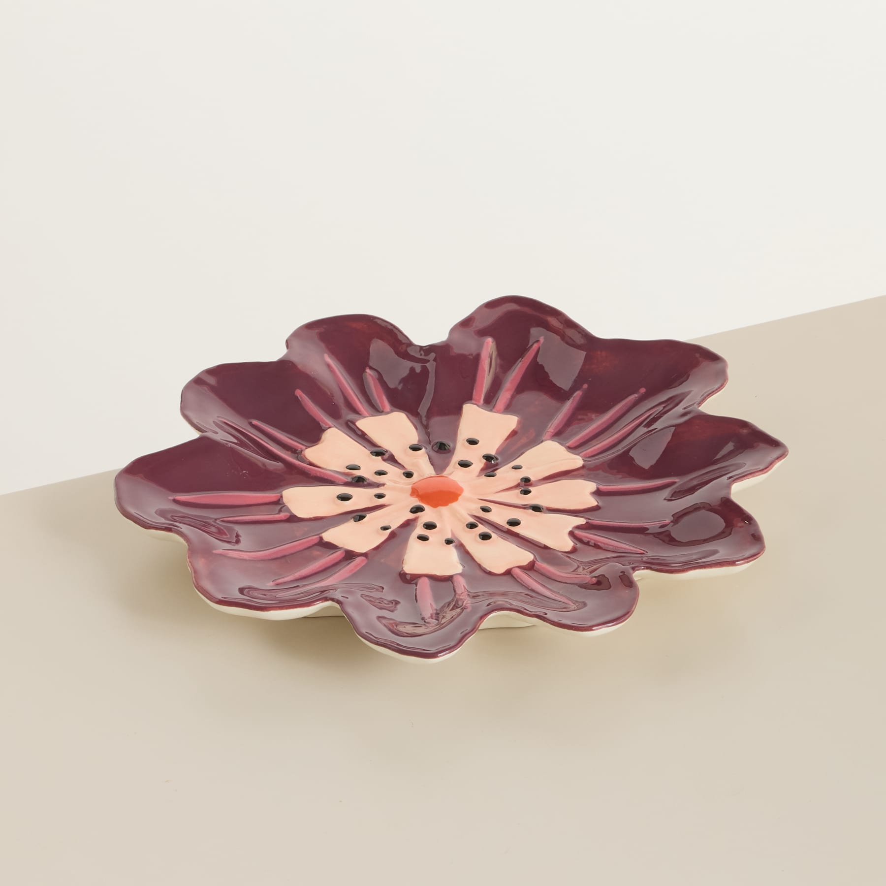 Eine leuchtende, burgunderfarbene Keramikblume mit zarten Blütenblättern und einem zentralen Staubgefäß sitzt auf einer schlichten, hellen Oberfläche.