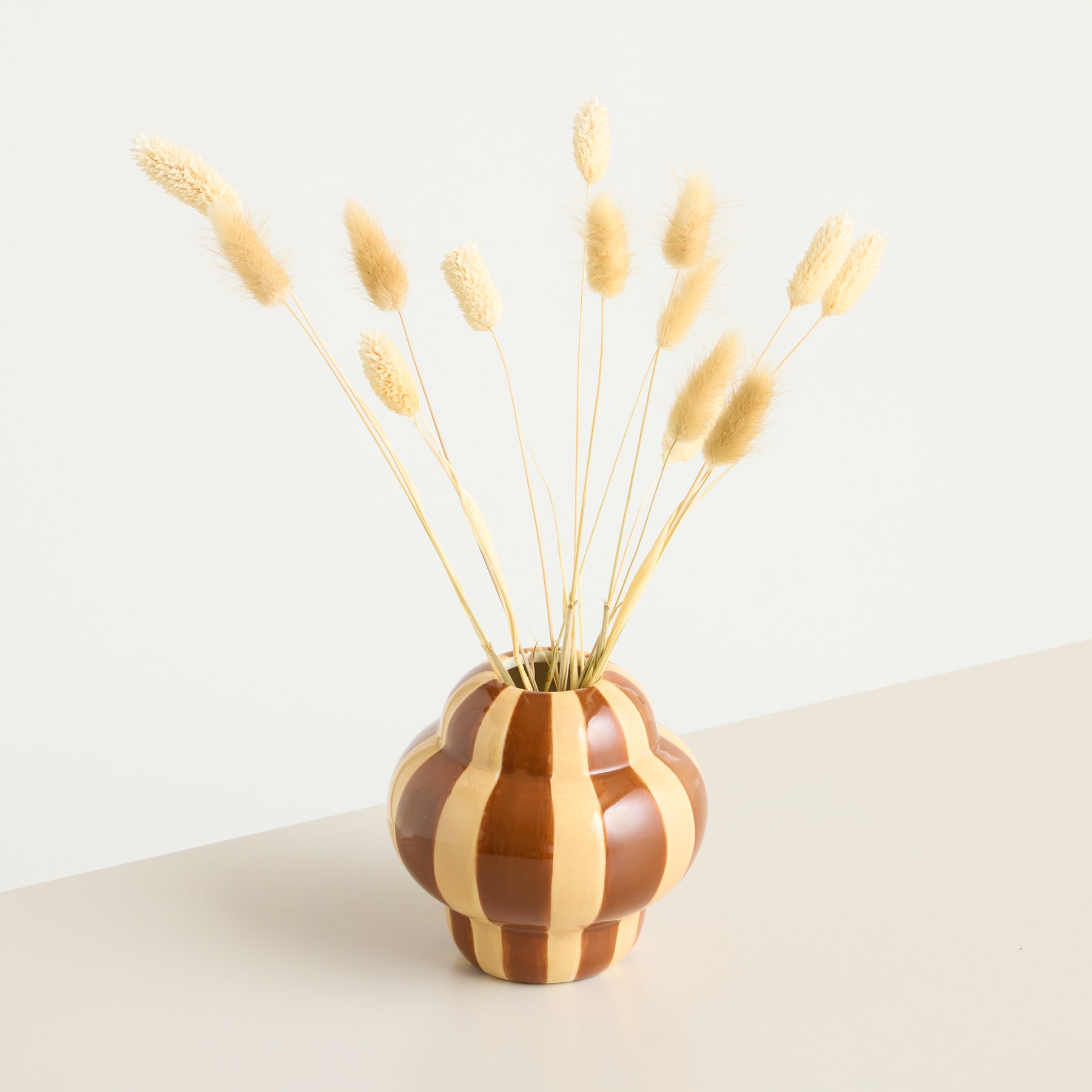 Eine dekorative Keramikvase mit Streifenmuster hält einen Strauß zarter, goldfarbener Weizenhalme vor einem schlichten, hellen Hintergrund.