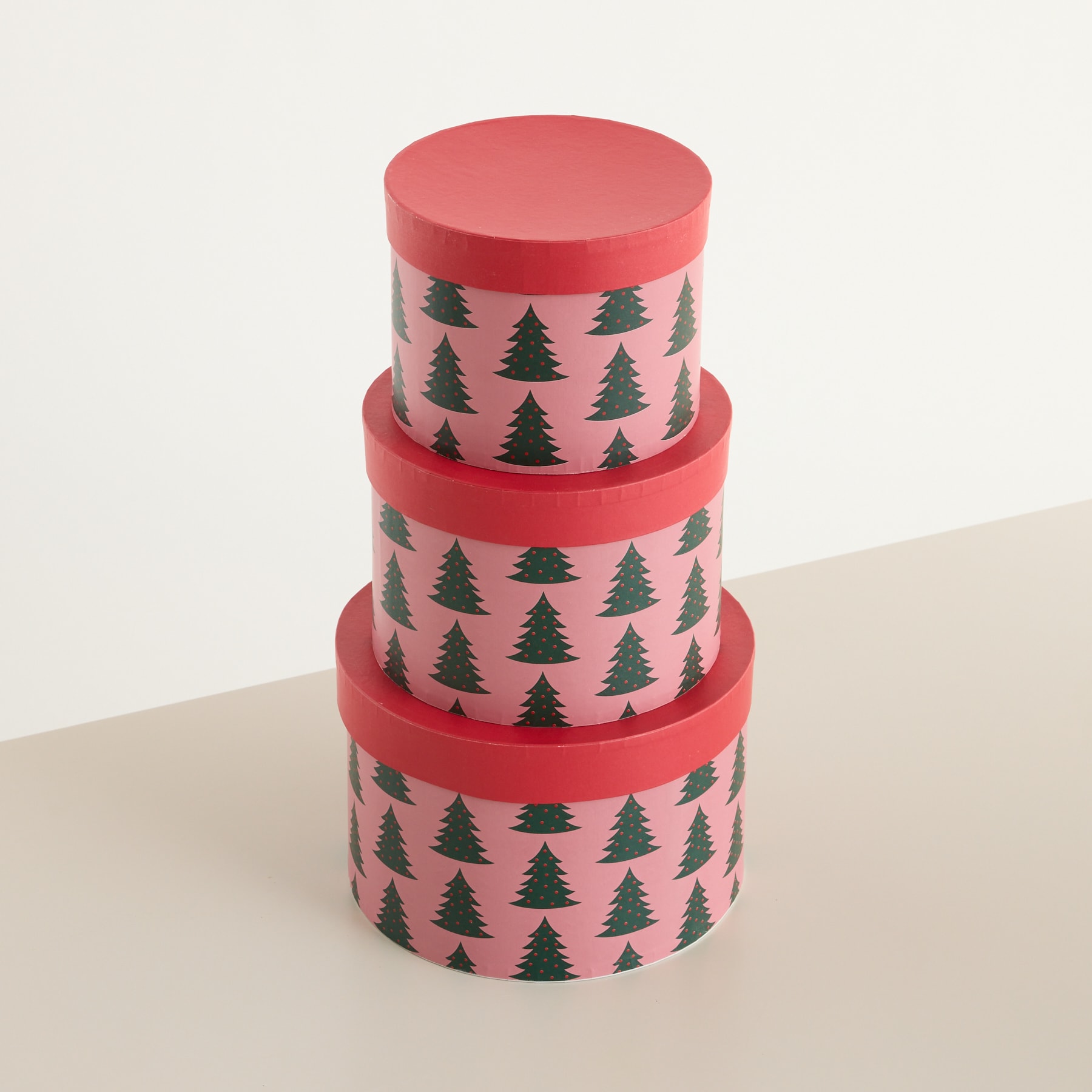 Ein Stapel aus drei runden, roten Geschenkboxen mit einem sich wiederholenden Muster aus grünen Weihnachtsbäumen darauf, auf einer hellen Oberfläche.
