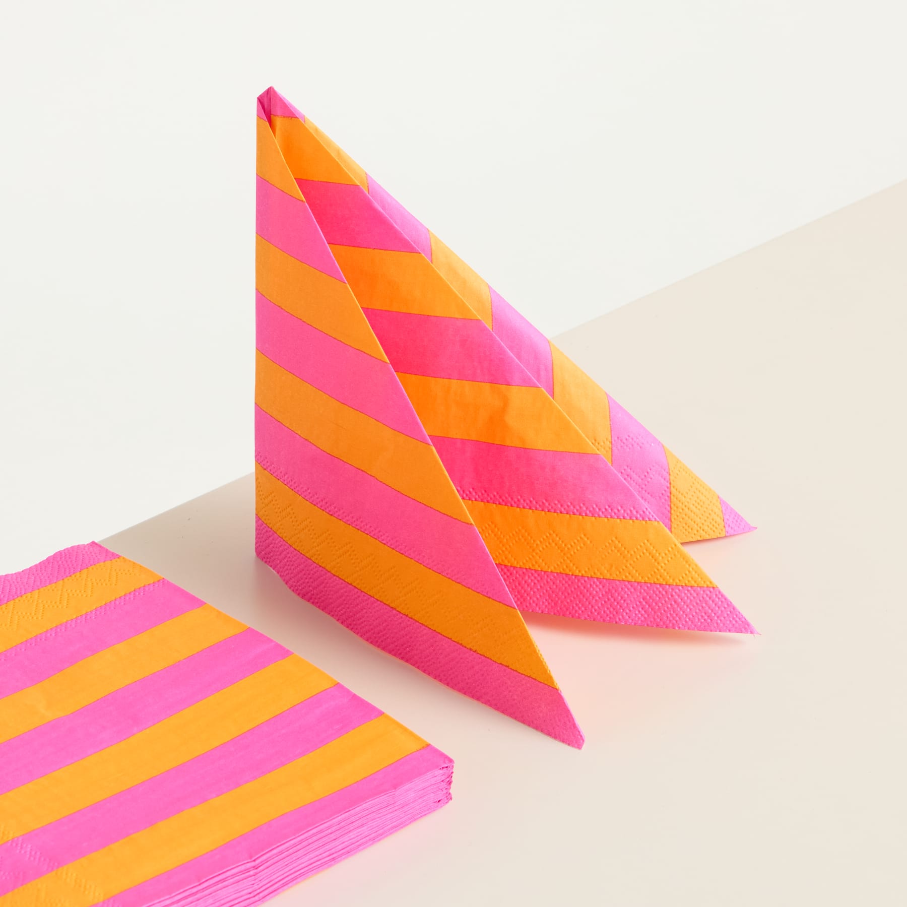 Eine lebendige, geometrische Papierskulptur mit kräftigen Streifenmustern in Rosa-, Orange- und Gelbtönen steht vor einem schlichten, hellen Hintergrund.