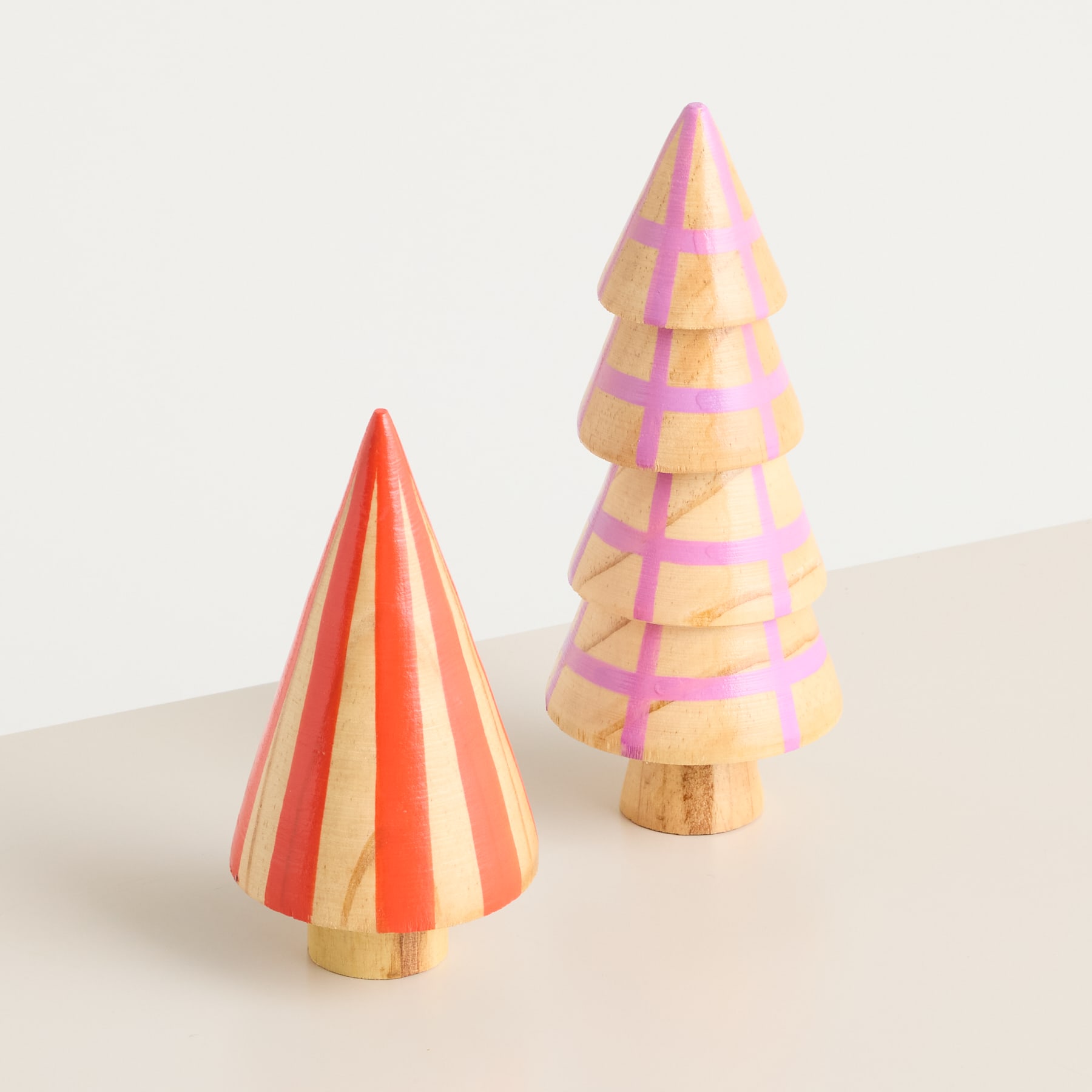 Zwei bunte Weihnachtsbäume aus Holz mit Streifenmuster stehen auf einer hellen Fläche vor einem schlichten Hintergrund.