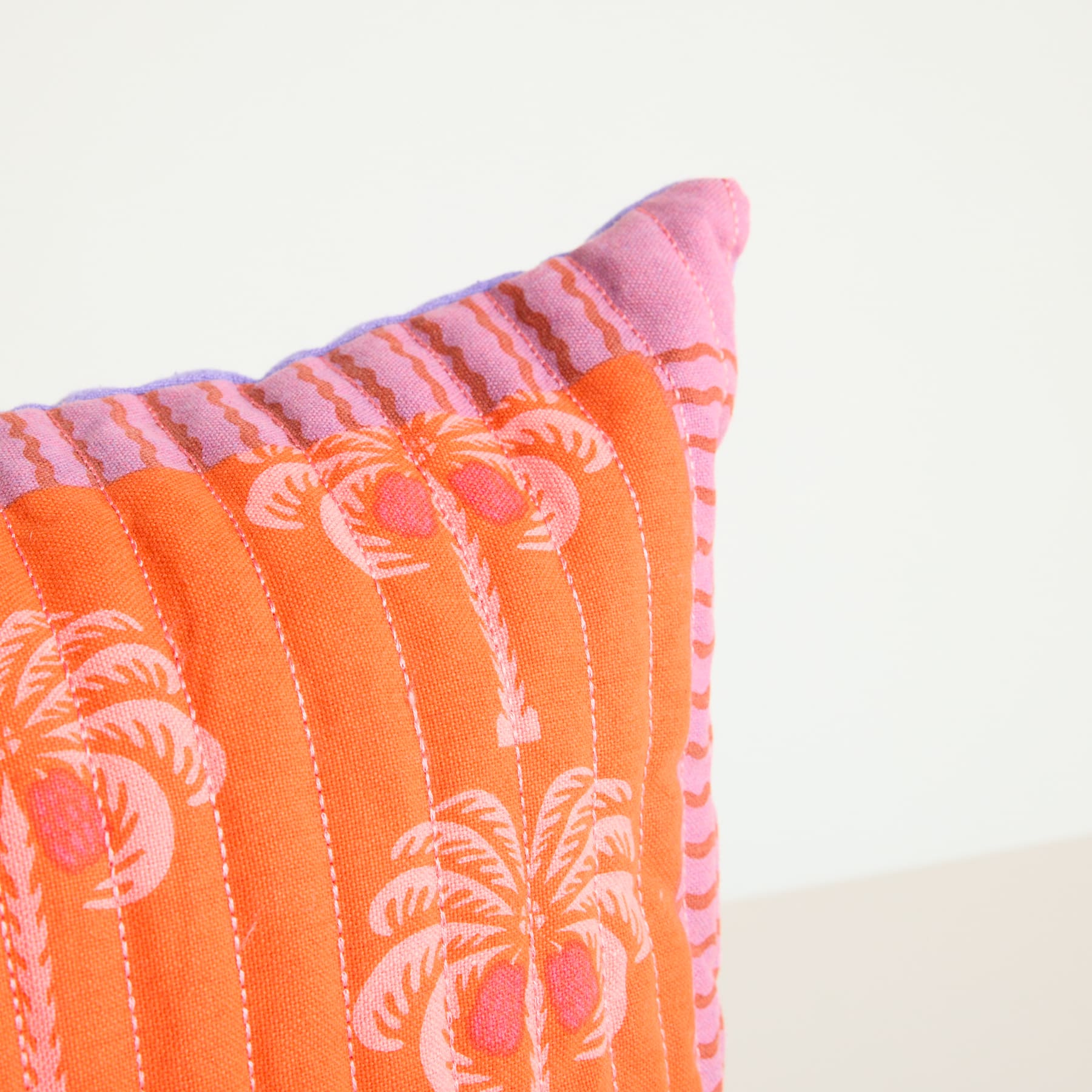 Ein farbenfrohes, gemustertes Kissen mit Palmenmotiven in Orange- und Rosatönen auf schlichtem Hintergrund.