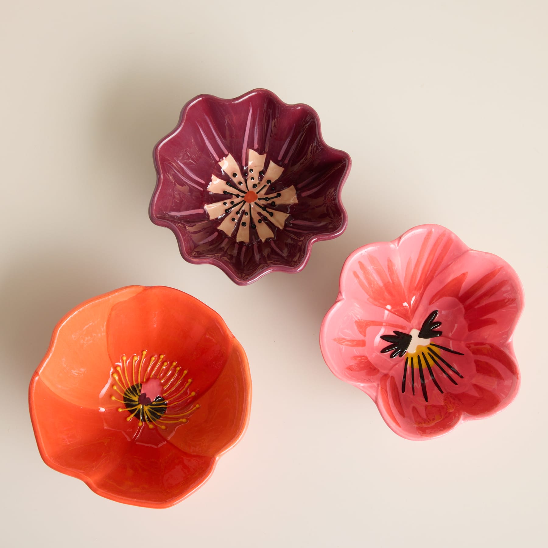 Drei farbenfrohe, blumenförmige Keramikschalen in Rot-, Orange- und Rosatönen werden vor einem neutralen beigen Hintergrund präsentiert.