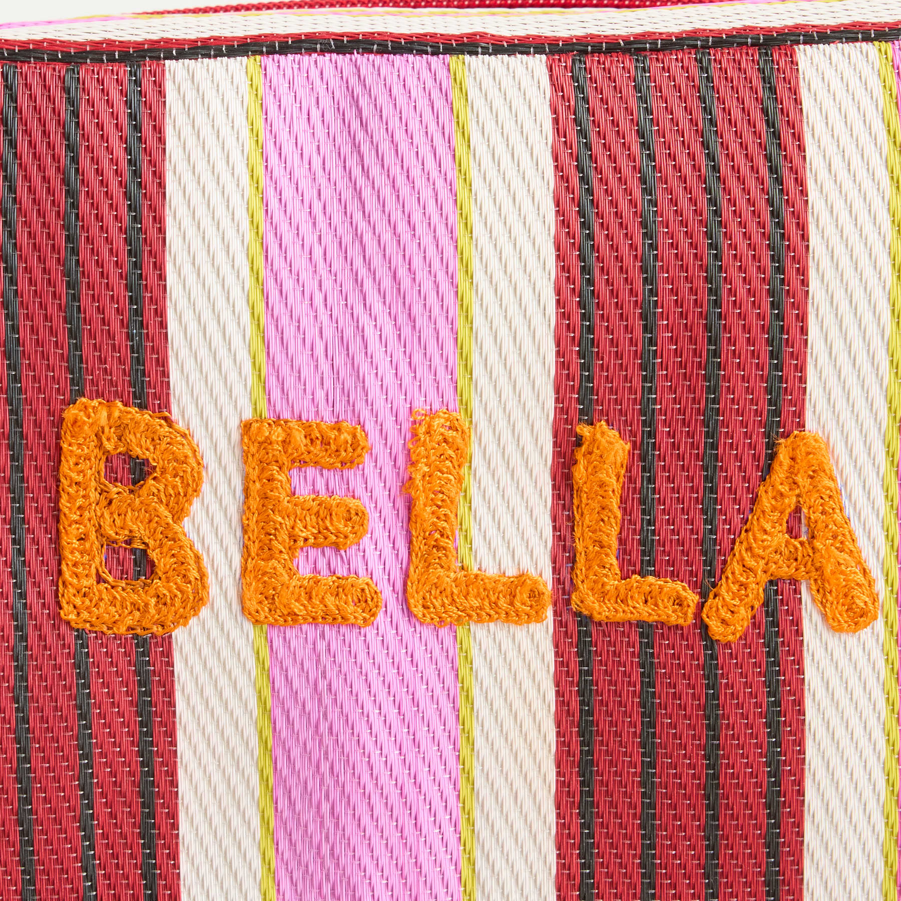 Das Bild zeigt das Wort „BELLA“ in großen, orangefarbenen, gehäkelten Buchstaben vor einem Hintergrund aus leuchtenden, mehrfarbigen Streifen.