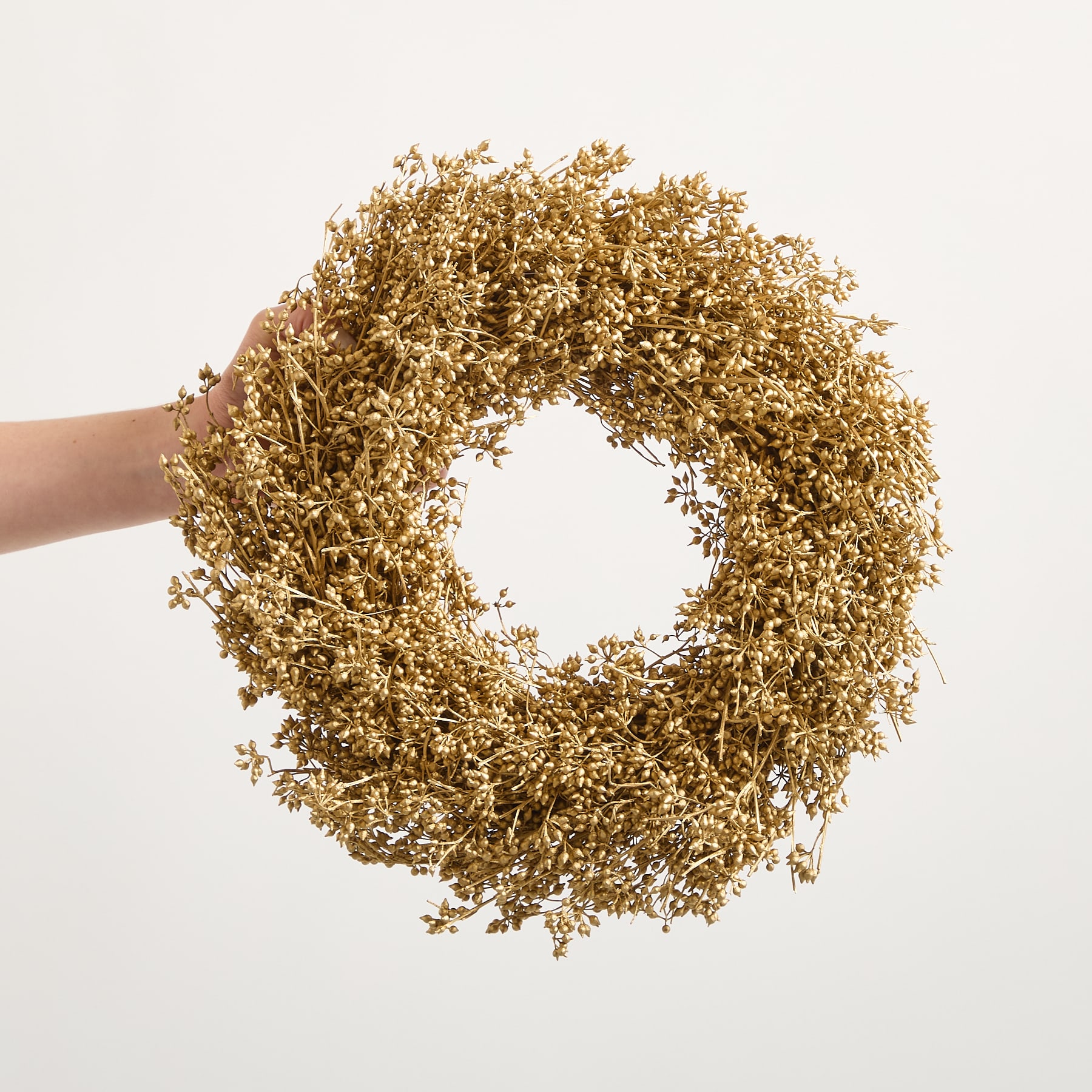 Eine goldene, kranzartige Dekoration aus getrockneten Blumen oder Blättern wird vor einem schlichten weißen Hintergrund in einer Hand gehalten.