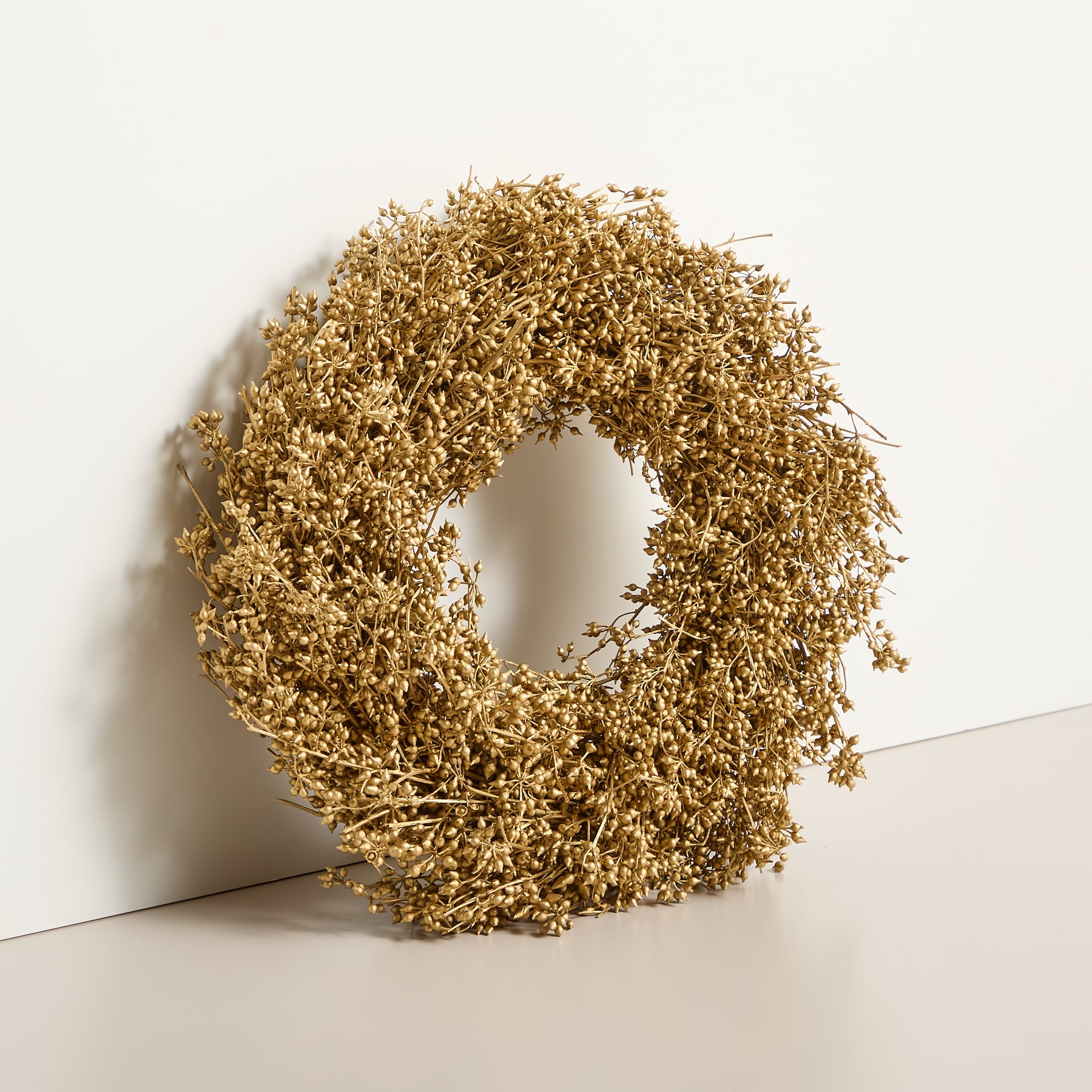 Auf einem schlichten weißen Hintergrund befindet sich ein goldener Kranz aus getrockneten Blumen oder Blättern.