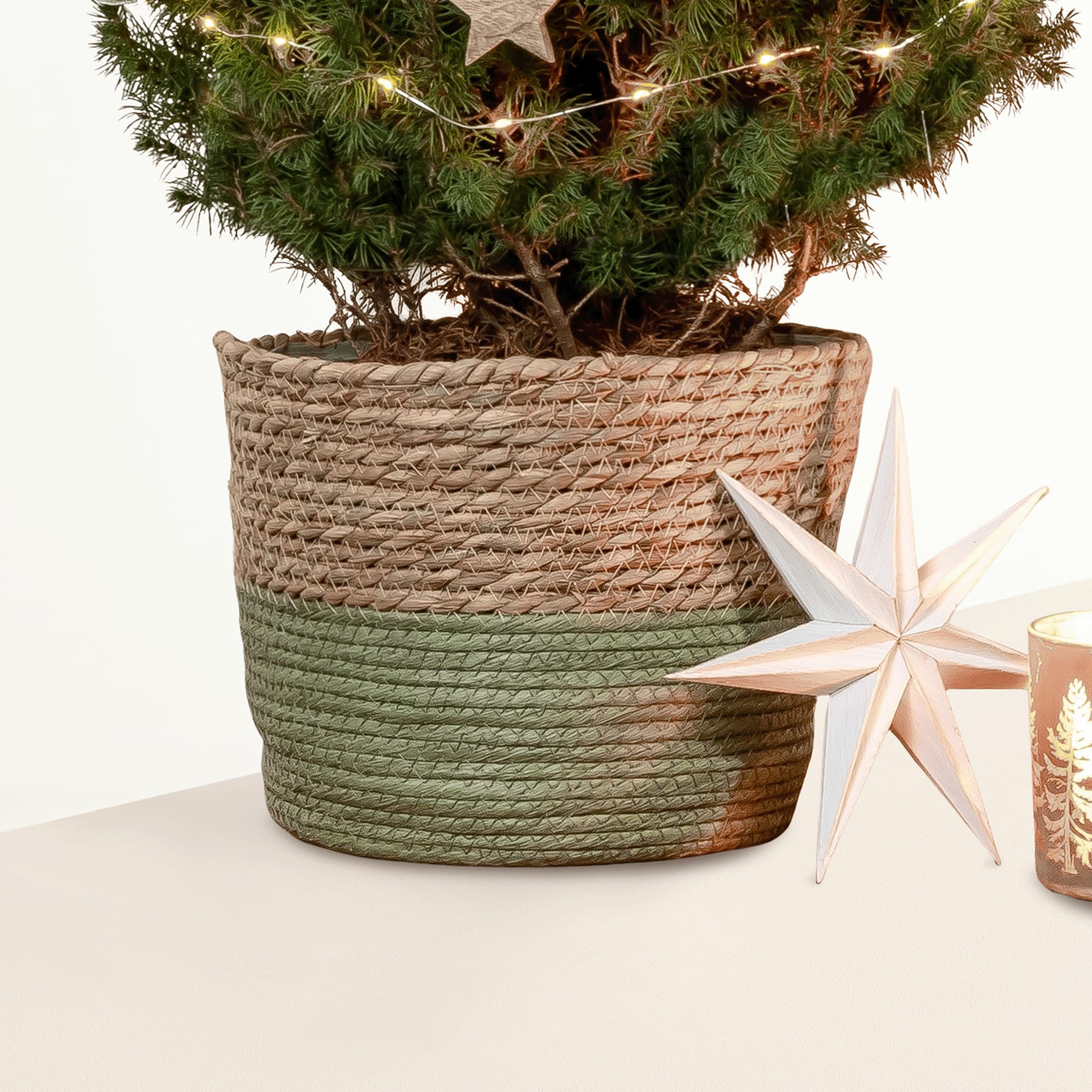 Eine gemütliche Weihnachtsszene mit einem geschmückten Baum in einem geflochtenen Korb, umgeben von festlichen Dekorationen, darunter ein sternförmiges Licht auf einem Holztisch.