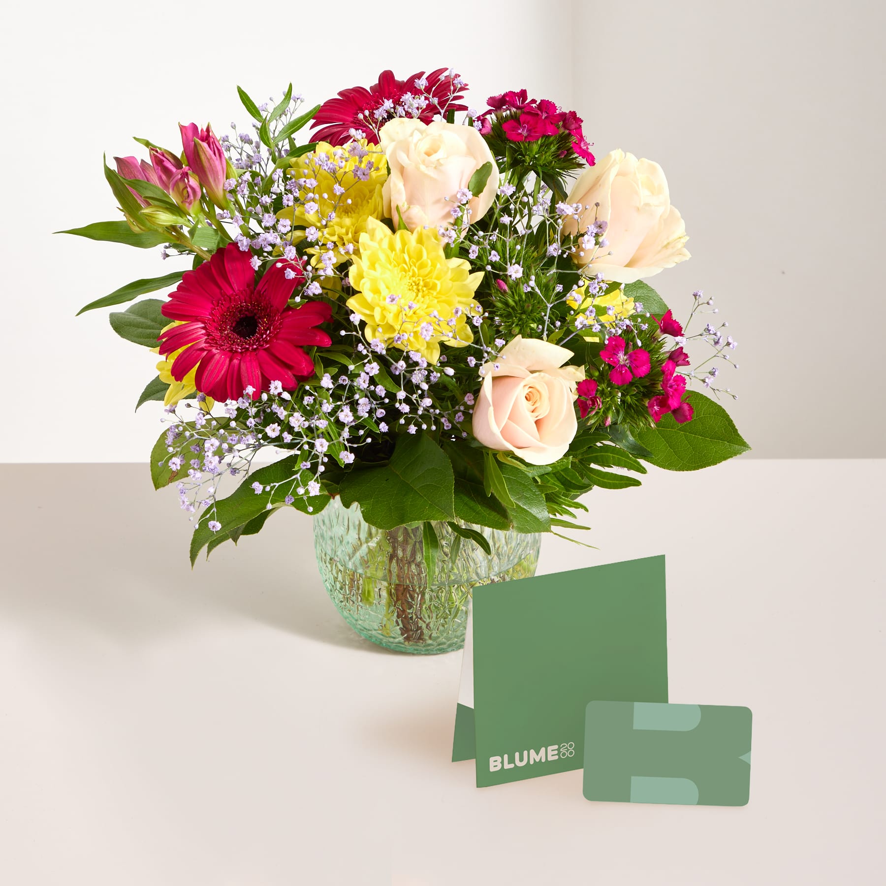 Ein lebendiger Strauß bunter Blumen, darunter Rosen, Gerbera und andere Blüten, wird in einer Glasvase auf einer Holzoberfläche präsentiert, begleitet von einer kleinen Grußkarte.