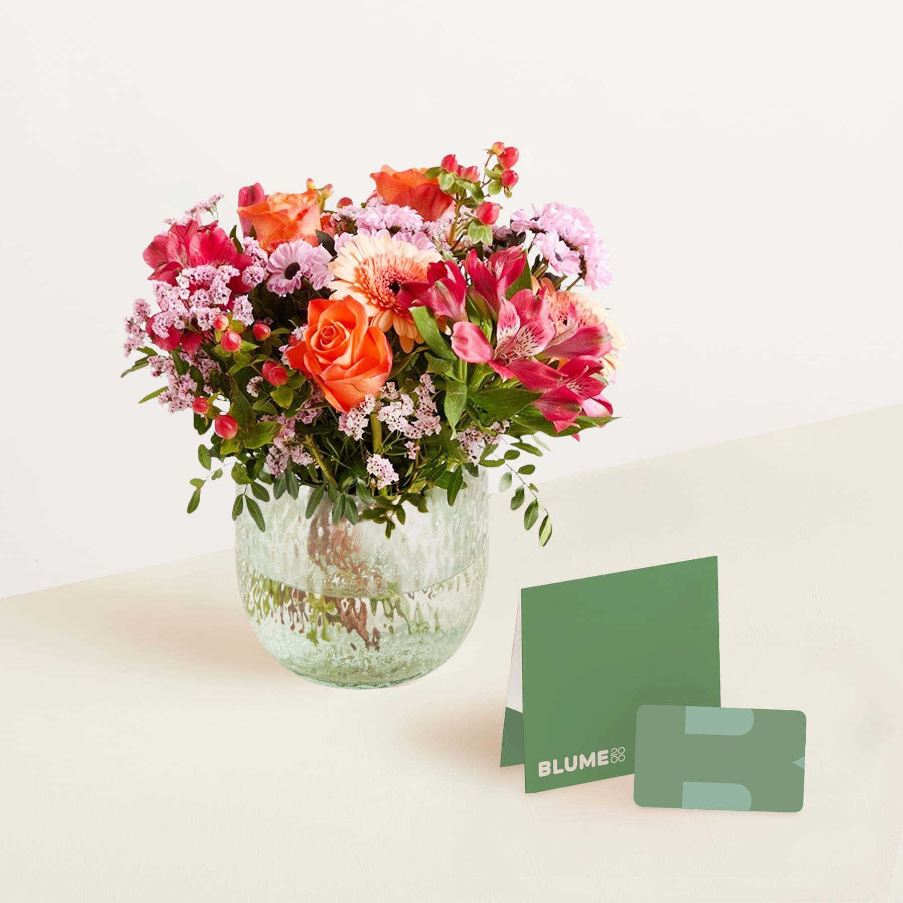 Ein lebendiger Strauß bunter Blumen, darunter Rosen und Nelken, wird in einer Glasvase auf einem schlichten Hintergrund präsentiert, begleitet von einer kleinen Geschenkbox.