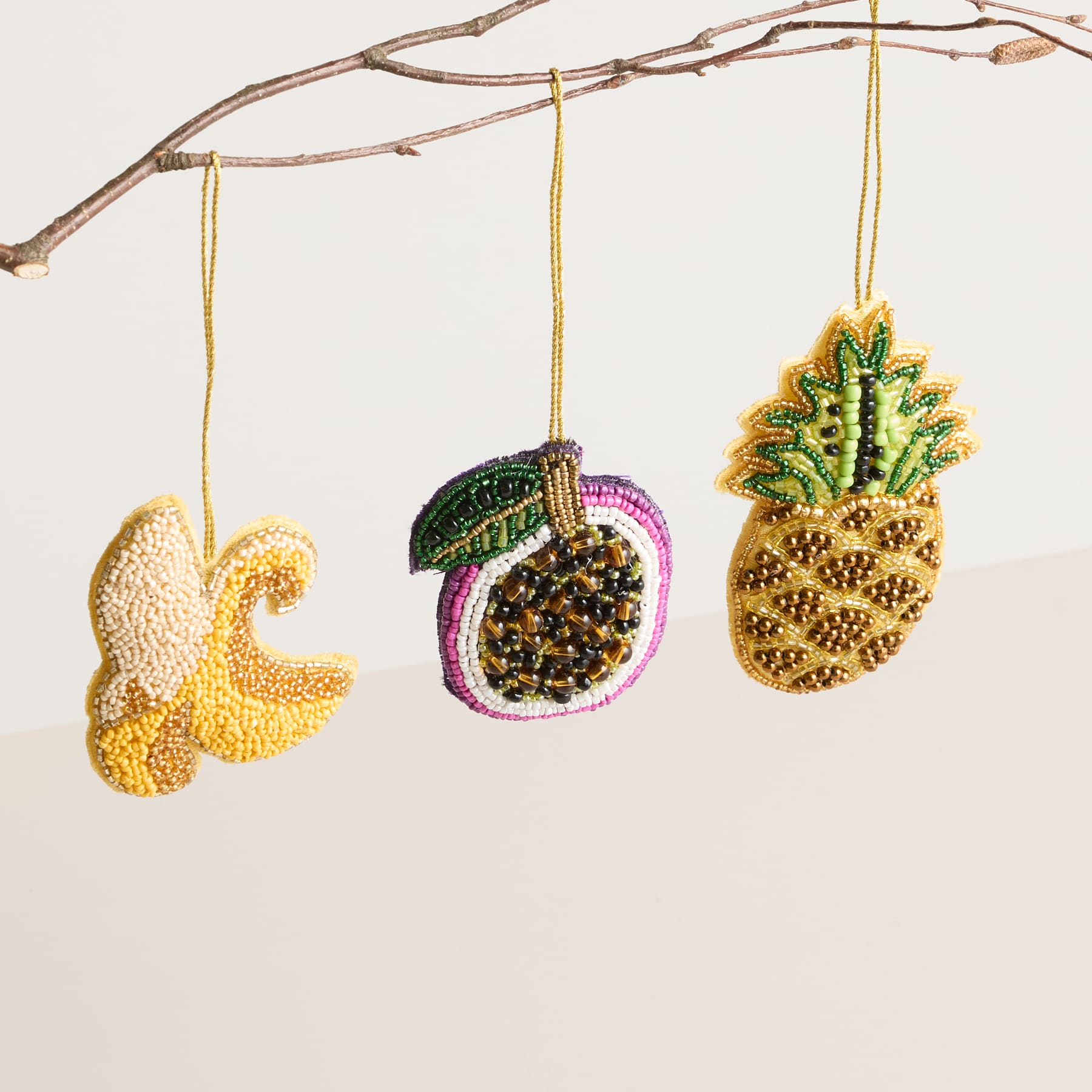 Bunte Perlenornamente in Form einer Banane, eines Apfels und einer Ananas hängen vor einem schlichten weißen Hintergrund von einem kahlen Ast.