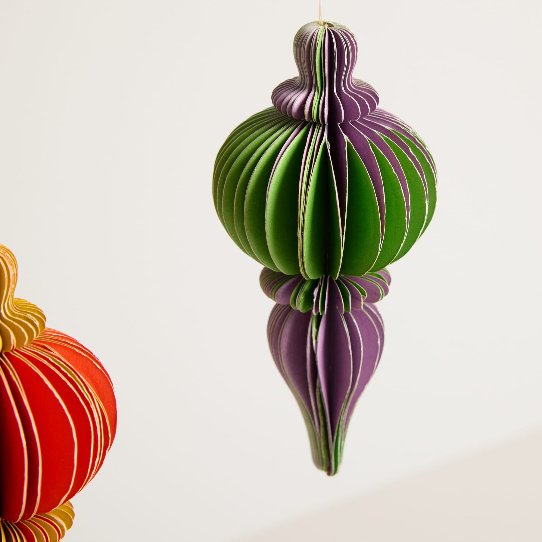 Ein lebendiges, origamiartiges Ornament in Grün- und Lilatönen hängt vor einem schlichten, hellen Hintergrund.