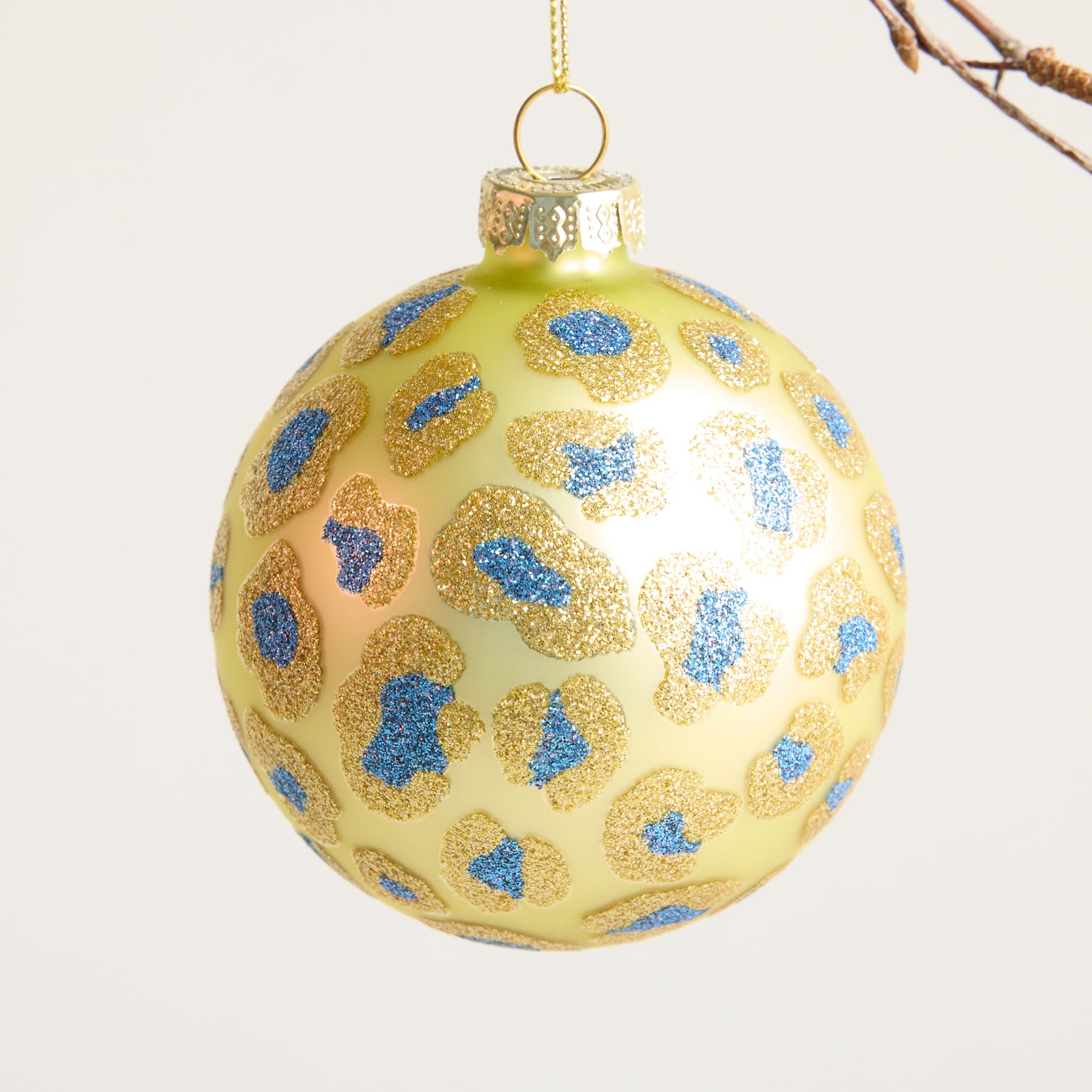 Ein goldenes Glasornament mit blau-weiß gesprenkelten Mustern hängt vor einem hellen Hintergrund an einem Ast.