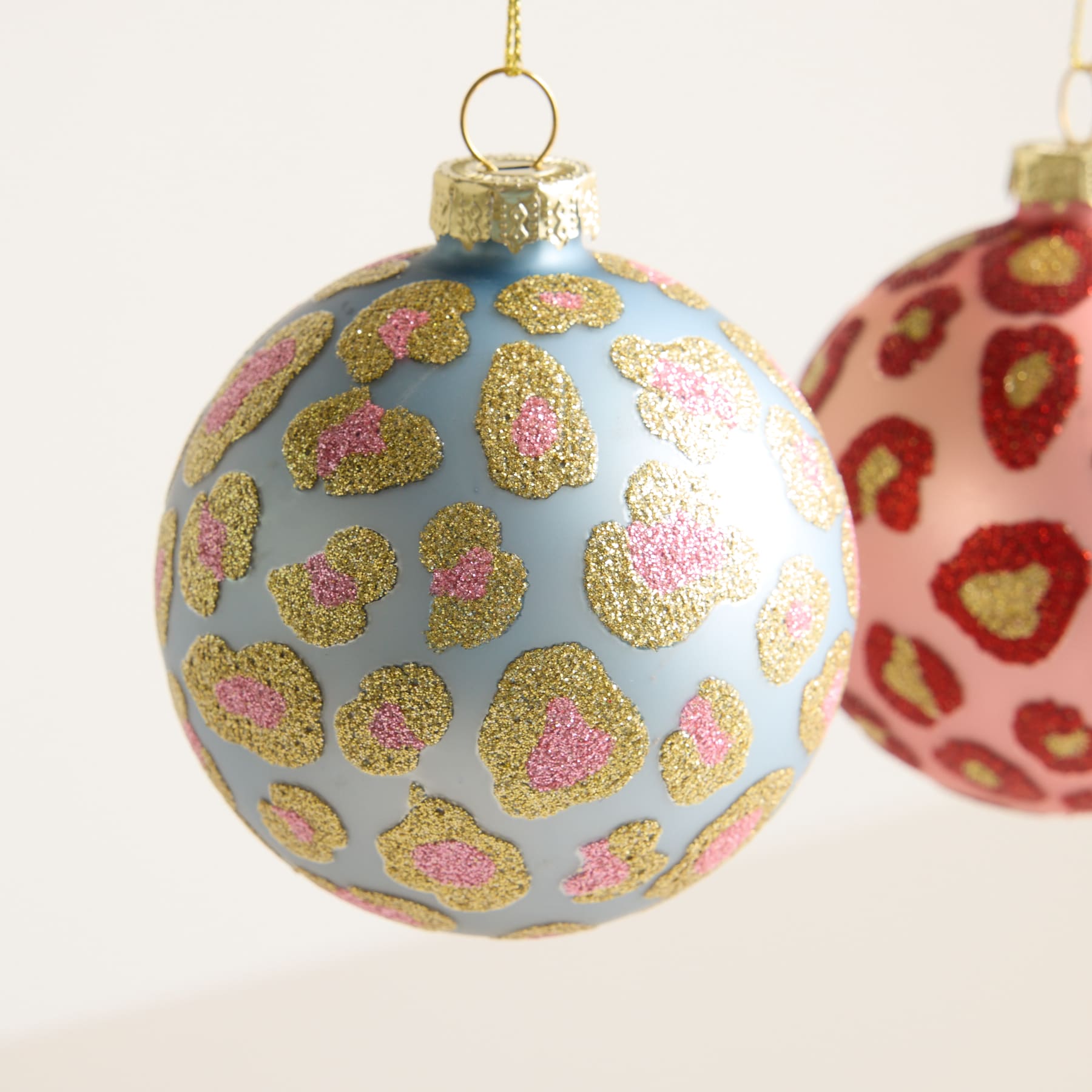 Das Bild zeigt ein Glasornament mit hellblauem Hintergrund und glitzernden Leopardenmustern in Gold und Rosa.