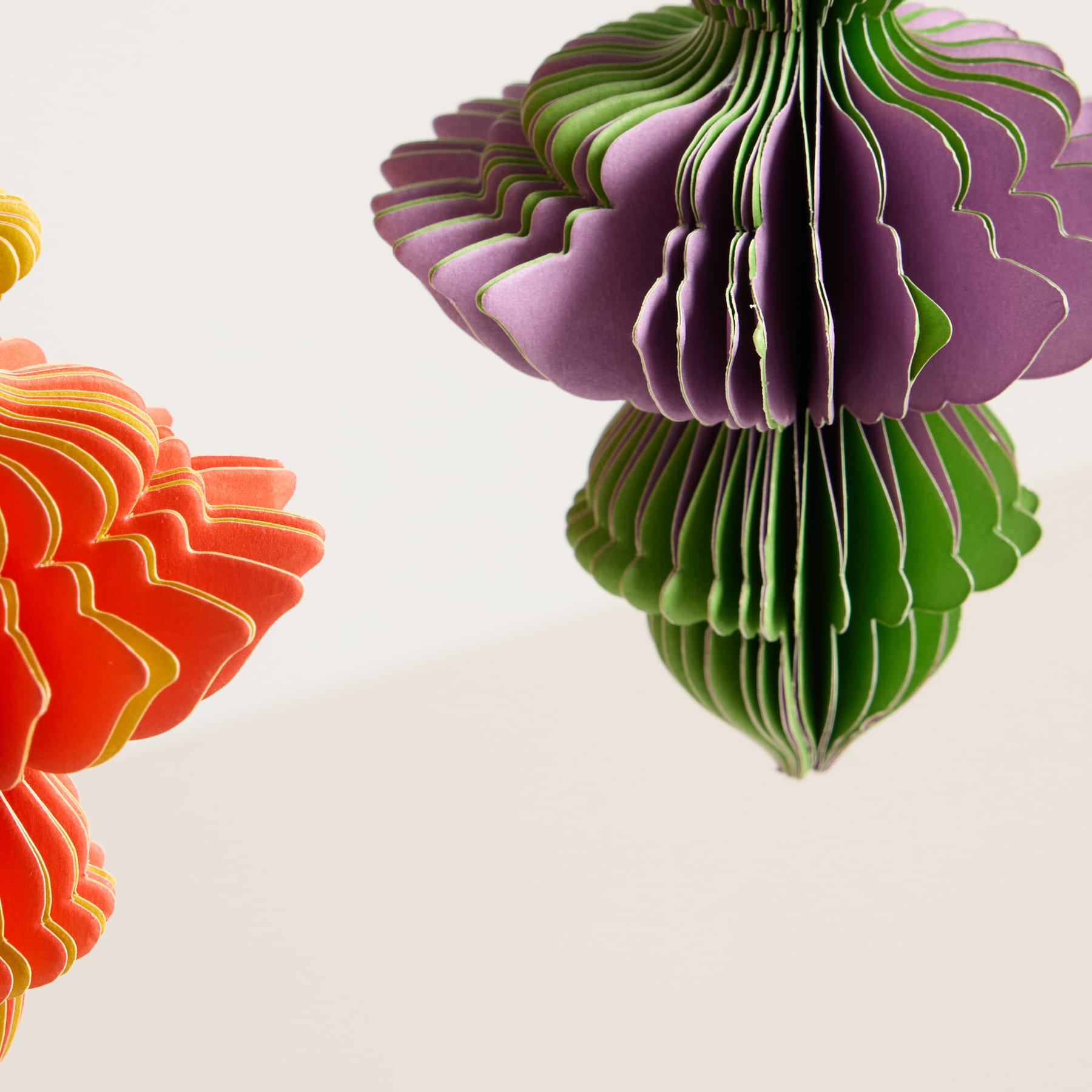 Lebendige, geschichtete Papierskulpturen in Grün-, Lila- und Orangetönen, die vor einem schlichten weißen Hintergrund eine auffällige und abstrakte, blumenartige Komposition bilden.