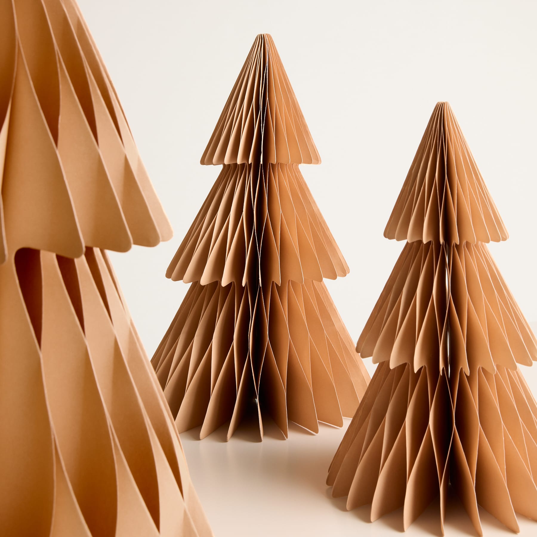 Aufwendige Papierskulpturen in verschiedenen Brauntönen, die stilisierten Weihnachtsbäumen ähneln, stehen vor einem schlichten weißen Hintergrund.