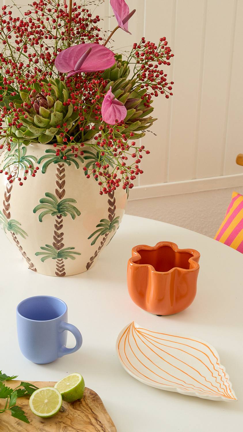 Auf einem Tisch steht ein farbenfrohes Blumenarrangement in einer gemusterten Keramikvase, dazu eine blaue Tasse, eine Limettenscheibe und eine orangefarbene Keramikschale.