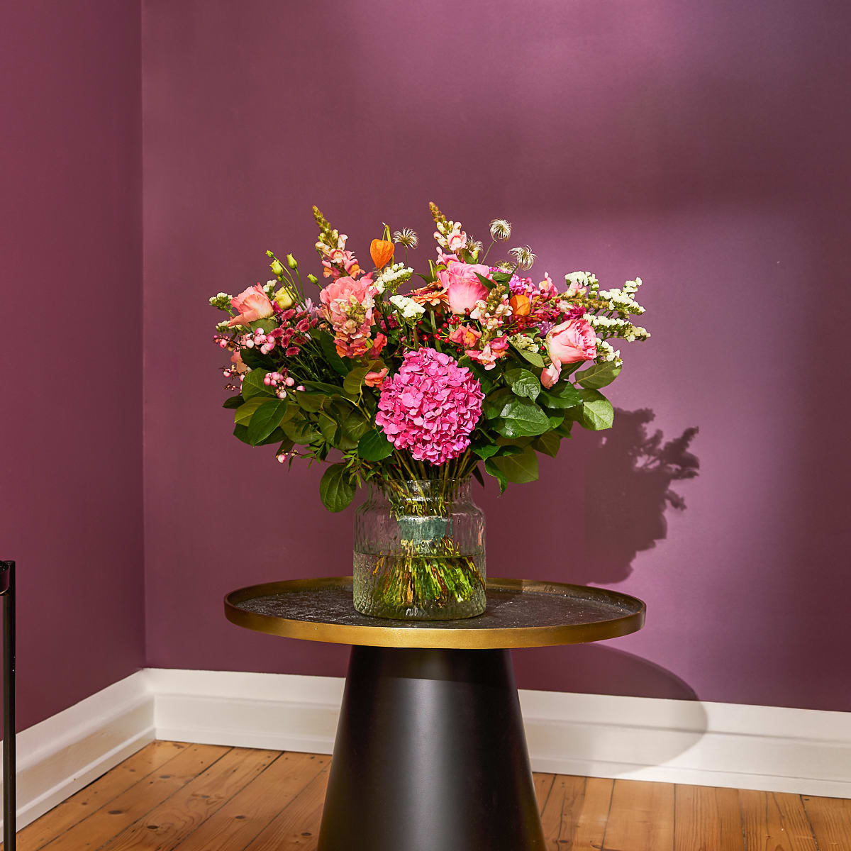 Ein farbenfroher Strauß rosa und gelber Blumen steht auf einem Holztisch vor einer dunkelvioletten Wand und schafft eine warme und einladende Atmosphäre.